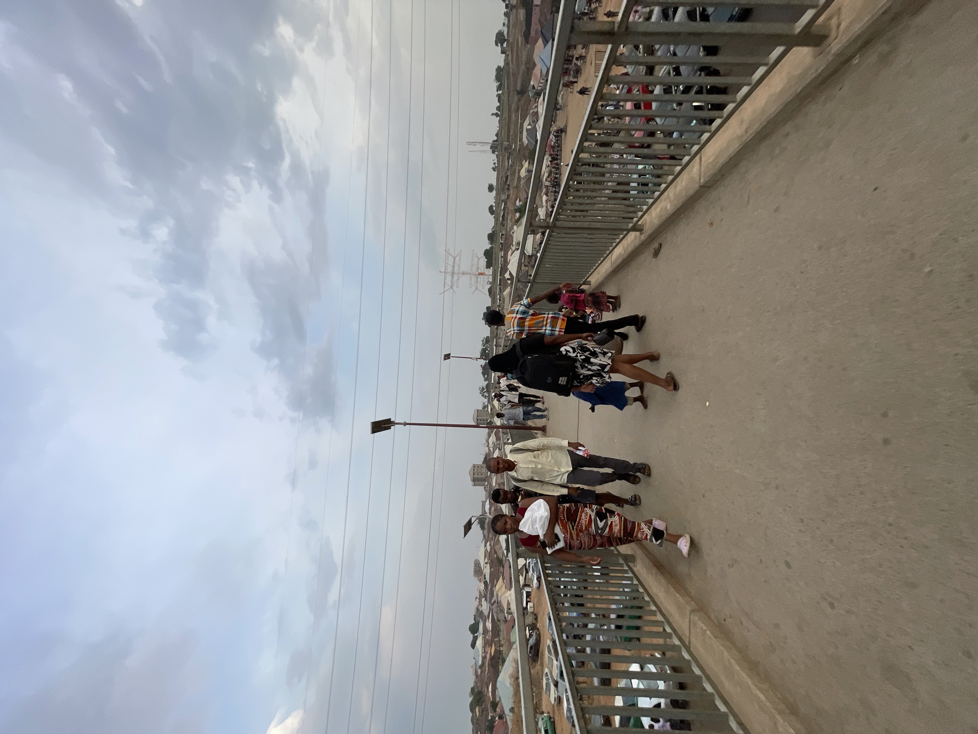 A long shot captures a group of pedestrians traversing a bridge in Abuja, Nigeria, under an overcast sky. The bridge, constructed of gray concrete and framed by metal railings, appears to be elevated above a dense urban landscape. A small group of people are walking across the bridge in both directions. A street lamp and power lines extend horizontally. The surrounding area shows a mix of low-rise buildings, and there's an indication of other vehicles.