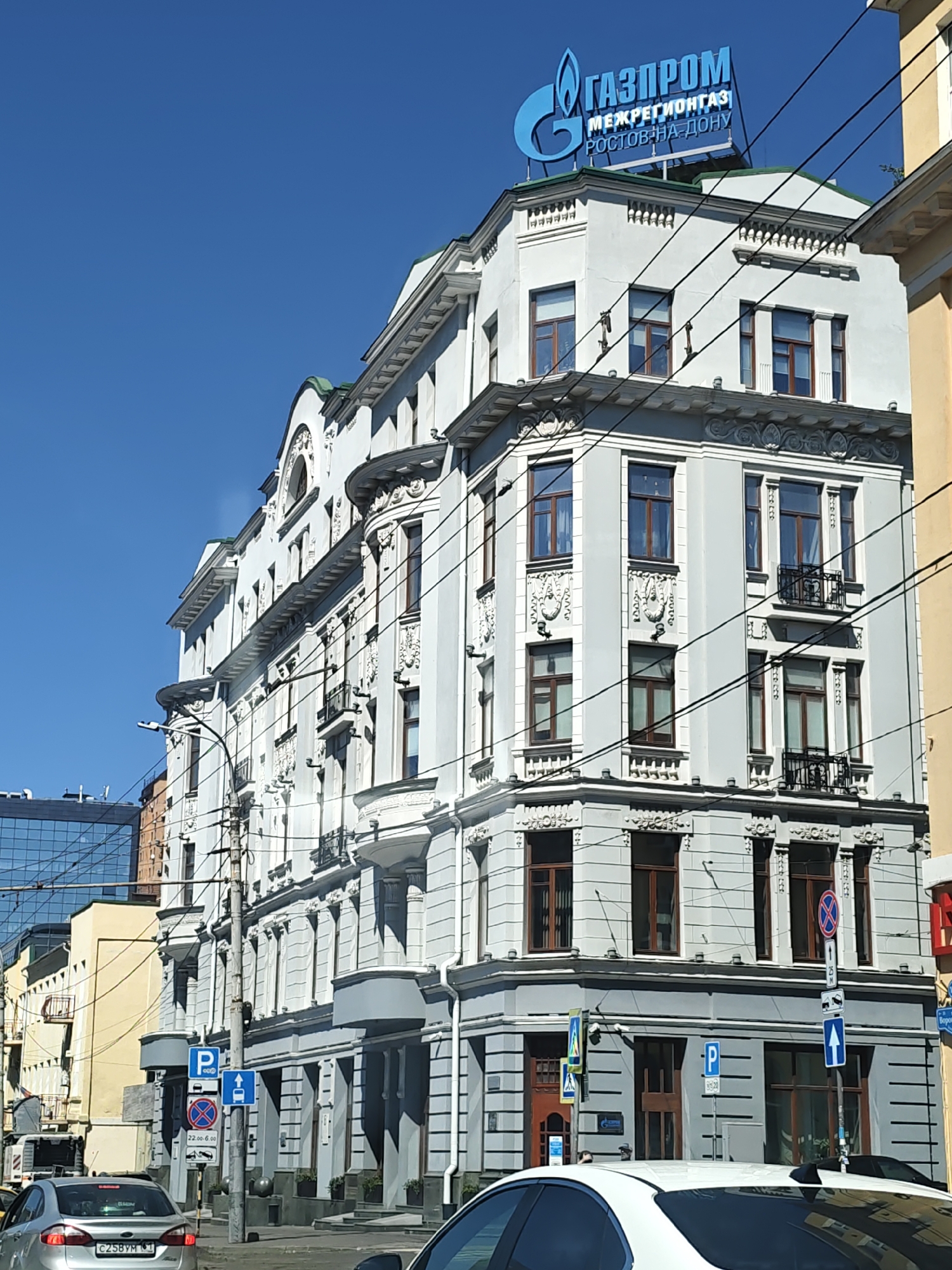 The image shows the Gazprom Mezregiongaz building in Rostov-on-Don, Russia. The building is a multi-story white structure with ornate architectural details, including columns, decorative carvings, and balconies. A large blue Gazprom logo is visible on the roof. The sky is clear and blue. In the foreground, there are cars on a street, and traffic signs are visible, including parking signs and a no-stopping sign. Power lines are strung across the scene.