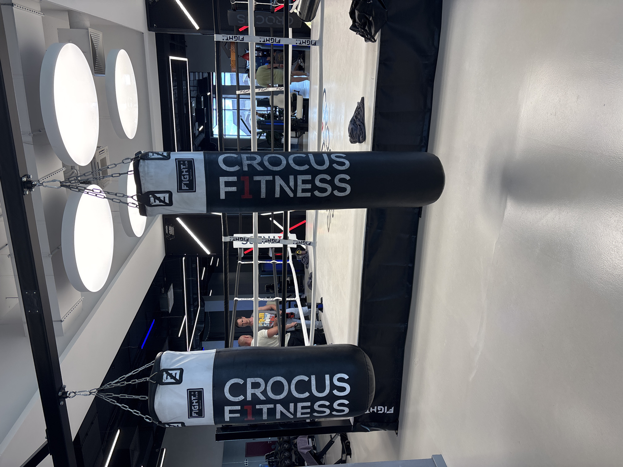 The image depicts the interior of a boxing gym, specifically the "Crocus Fitness" facility in Monolit, Russia. In the foreground, two large punching bags hang from chains, one white and black, the other black. The black punching bag has the "Crocus Fitness" logo printed vertically in white. A boxing ring is visible behind the punching bags, with its ropes and padded corners. Two men are standing inside the ring, appearing to converse. The gym's ceiling features modern circular lighting fixtures and a black grid system. The floor is a light gray, with some equipment and bags visible in the background. The overall environment suggests a professional and well-equipped fitness center.