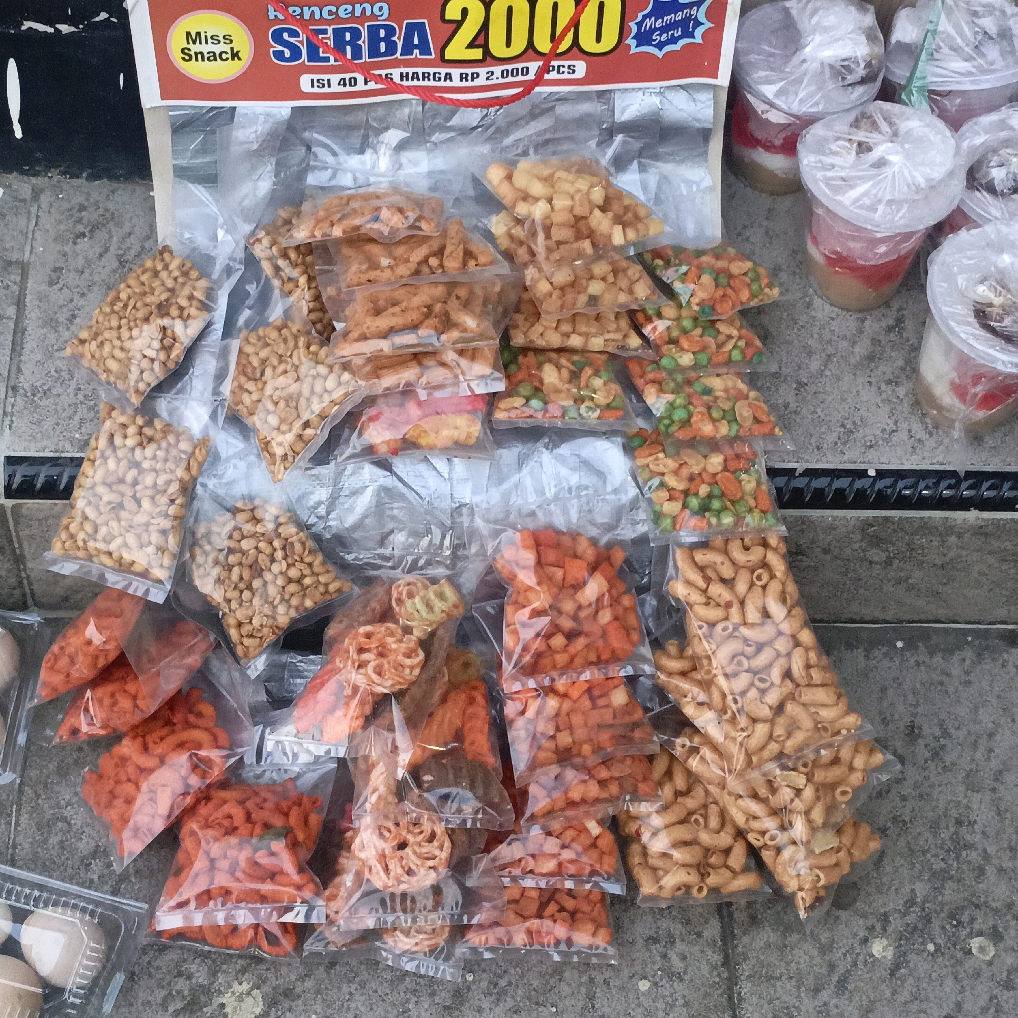 This is a high-angle, medium shot image of a street vendor's stall in Salatiga, Indonesia. The stall displays a variety of individually packaged snacks, primarily nuts and fried savory items, for sale. The snacks are arranged in clear plastic bags and some small plastic cups. The background shows a concrete surface and what appears to be a black grate or metal bar. The overall scene depicts a typical roadside commerce setup.
