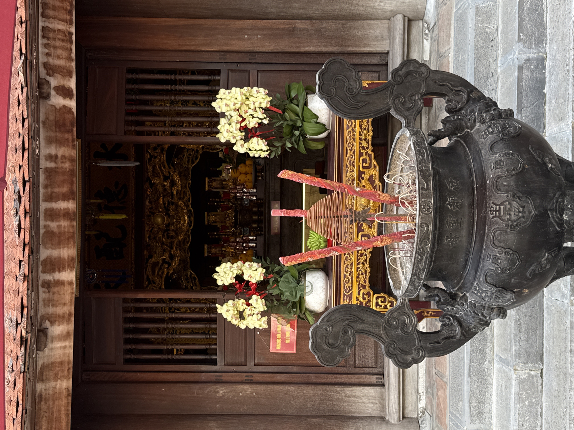 The image depicts a temple entrance in Hà Nội, Vietnam. The temple's wooden facade is visible, with intricate carvings and decorations. Two planters with white orchids flank the doorway. An ornate metal incense burner stands prominently in the foreground. It contains red incense sticks and coiled incense. The background shows the temple's interior, with religious artifacts and offerings. A stone staircase is to the right of the burner.