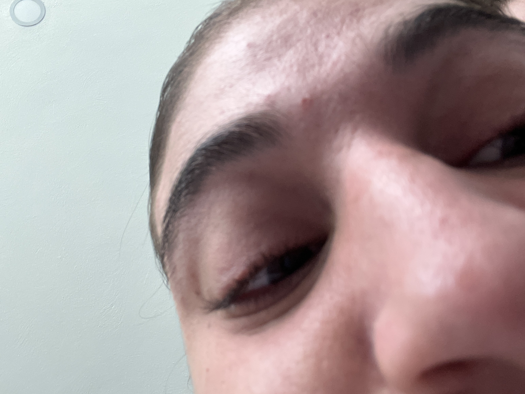 Close-up, partially obscured view of a person's face. The focus is primarily on the upper part of the face, featuring the forehead, eyebrow, eye, and part of the nose. The person has dark eyebrows and eyelashes. A red spot or blemish is visible on the forehead. The background is a plain, light-colored wall. There is a round device at the top left. There is no evidence to place the scene in Puebla, Mexico.