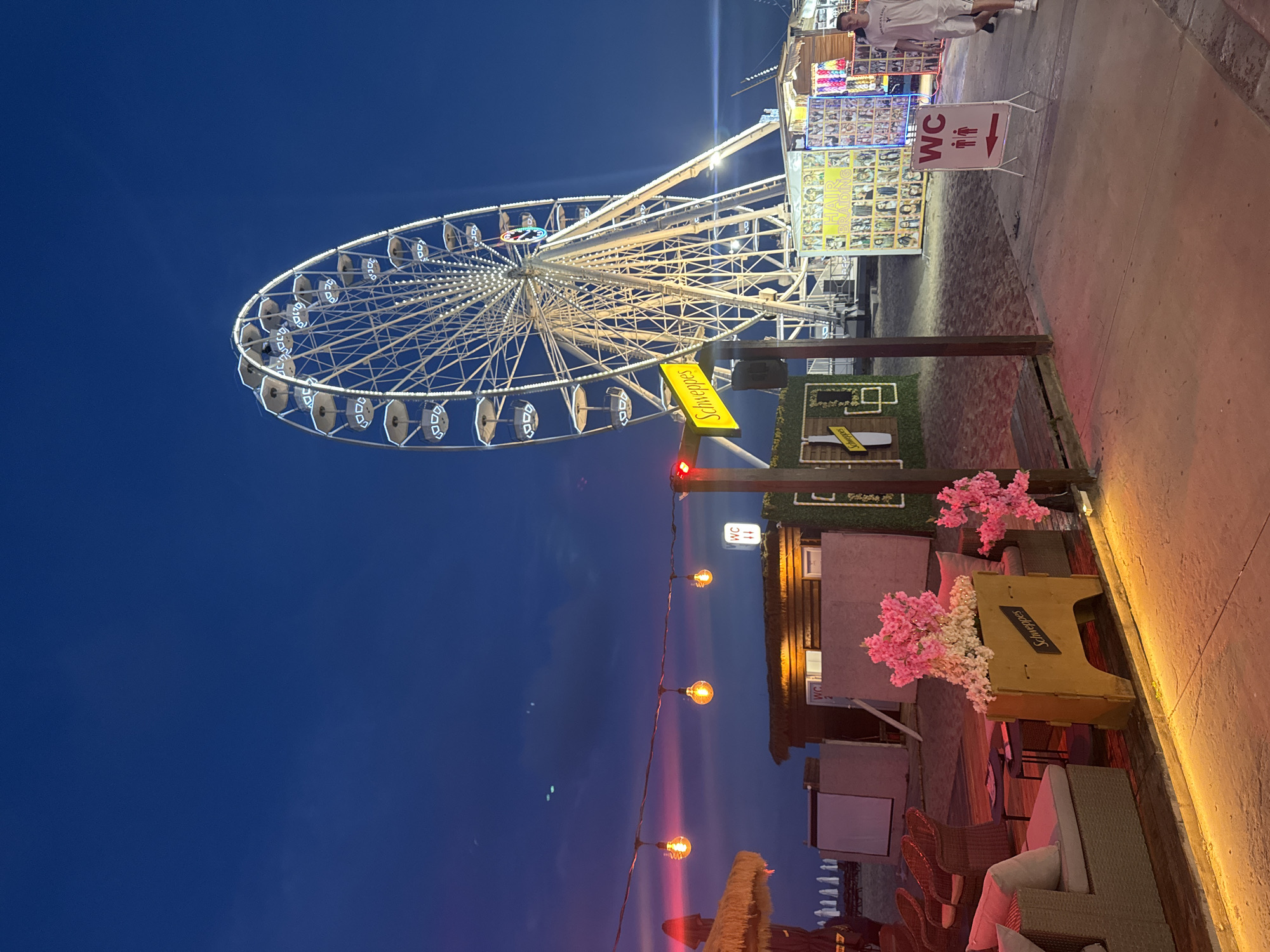 An outdoor scene at dusk features a large Ferris wheel illuminated with white lights against a dark blue sky. In the foreground, a beach area is set up with lounge seating, a small table, and decorative pink flowers in a planter. String lights with glowing orange bulbs hang above a rustic wooden structure. To the right of the Ferris wheel, a sign for "Schweppes" is visible, and further to the right, stalls with various merchandise, including a "Hair Braiding" display, are present. A "WC" sign with directional arrows is also visible. A man in a white t-shirt and shorts walks past the stalls. The ground is a mix of sand and concrete.