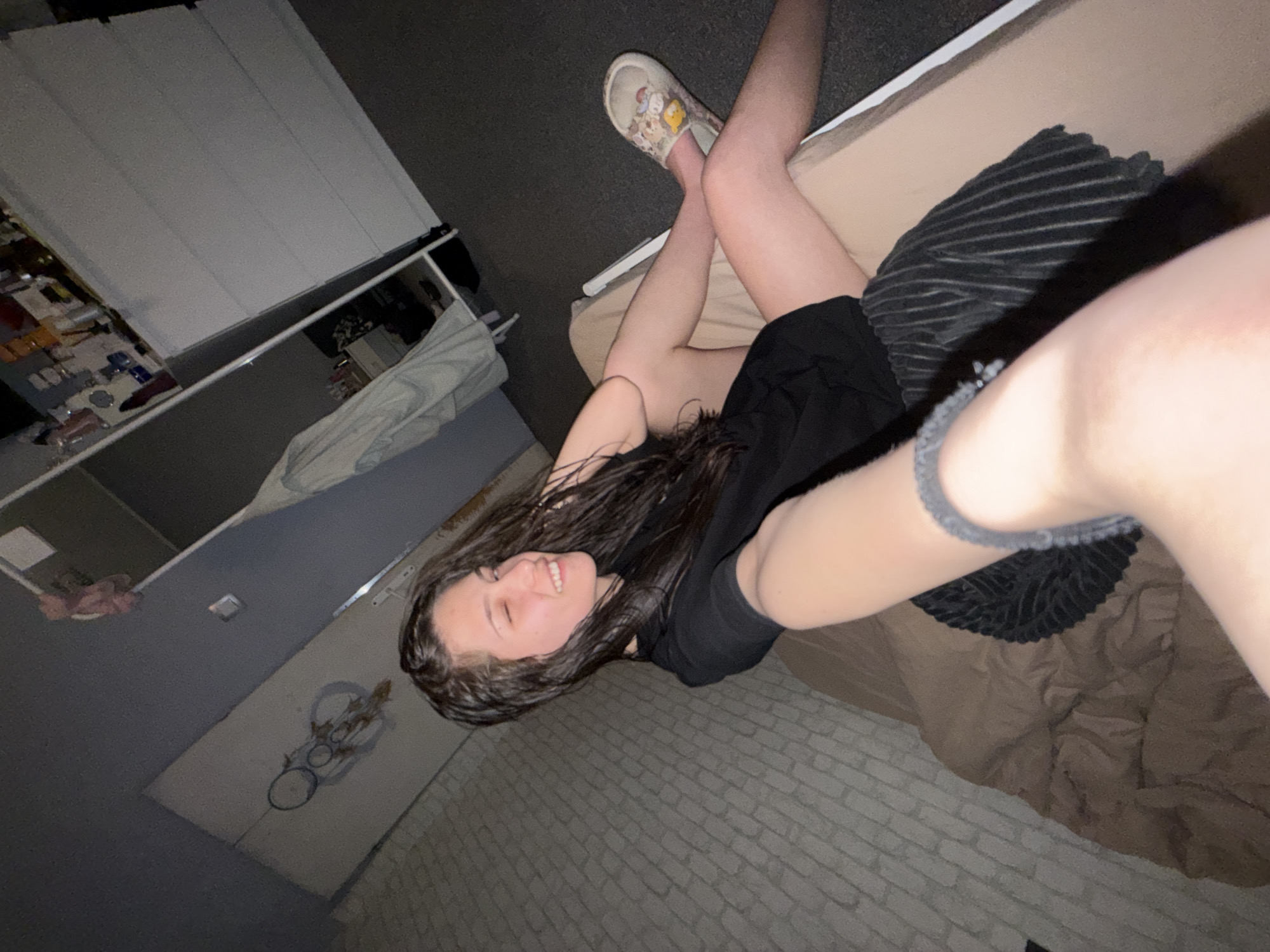 A woman with wet dark hair is lying on a bed, wearing a black shirt. Her left leg is crossed over her right, with a patterned shoe on her right foot. The bed has a tan comforter and a striped pillow. The room has a white closet or shelving unit, and a wall with a light-colored brick pattern. The image appears to be taken from a selfie perspective. Location is unknown.