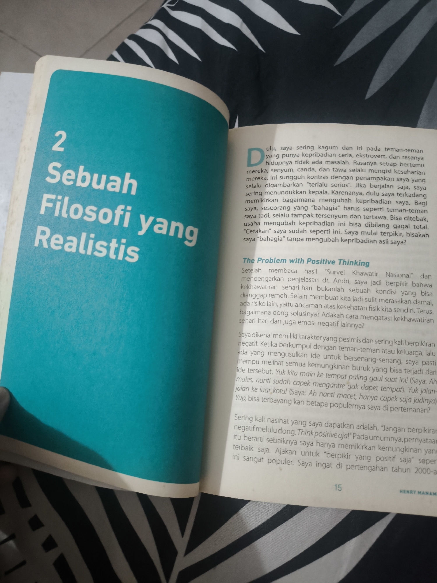The image is a close-up shot of an open book. The left page features a large, teal-colored title section with the number "2" followed by "Sebuah", "Filosofi yang", and "Realistis" printed in large white sans-serif font. The right page contains printed text in Indonesian. The text discusses the author's observations of friends with cheerful personalities and contrasts this with their own perceived seriousness. It also delves into the concept of positive thinking and the author's personal reflections on it, citing a "Survei Khawatir Nasional" and Dr. Andri. The bottom of the right page shows the page number "15" and the author's name "HENRY MANAM". The book is placed on a surface with a black and white striped pattern, likely fabric. The lighting suggests an indoor environment, possibly a room in Gentan, Indonesia, where the book is being read. There are no people or direct interactions depicted, only the book as the central subject.