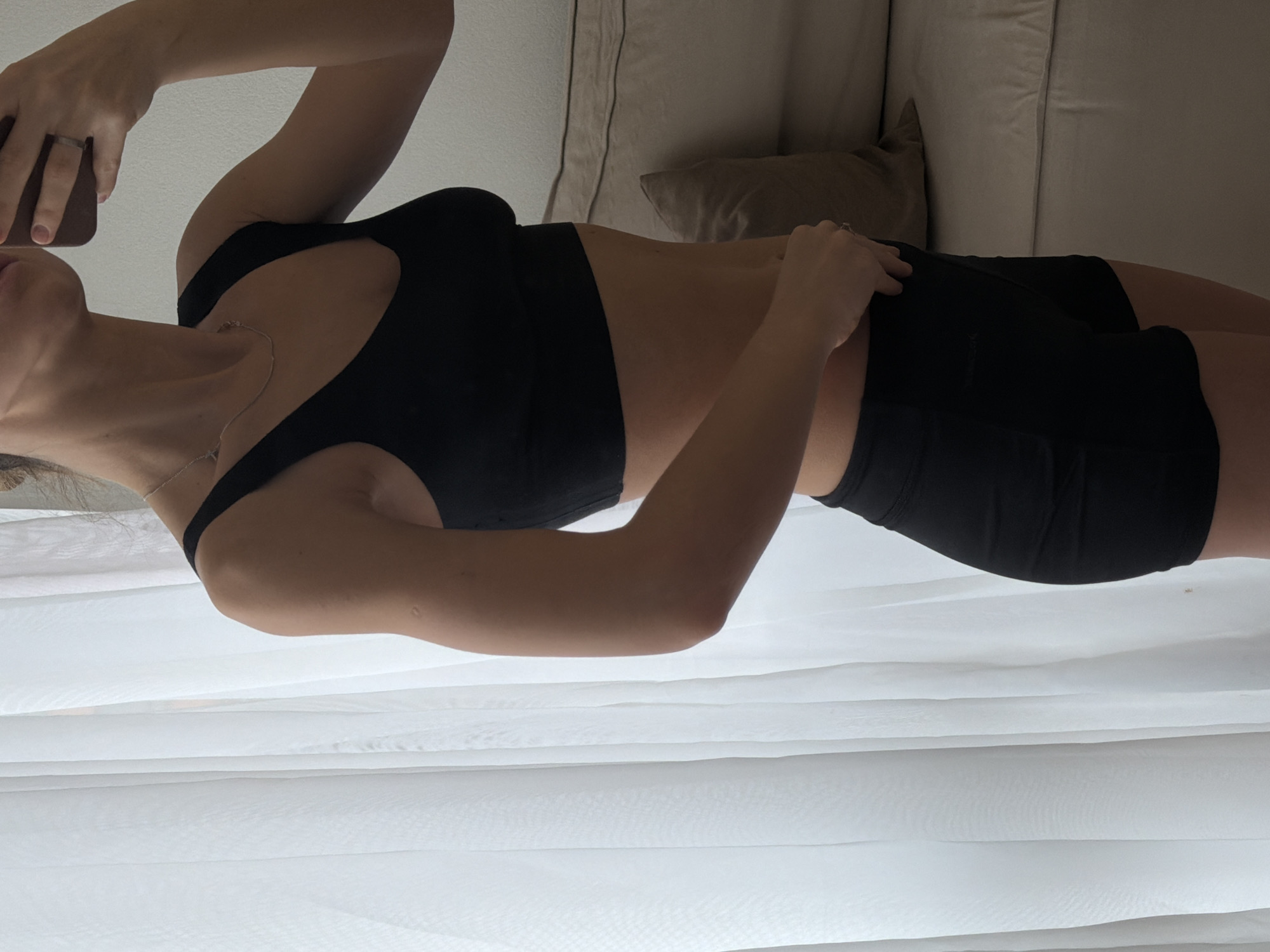 A woman lies on a white bed or couch, dressed in a black sports bra and black shorts. She holds a phone up and is likely taking a selfie. The scene includes a pillow behind her and part of the couch. There is no information in the picture to determine the location in Khimki, Russia, or provide any forensic details.