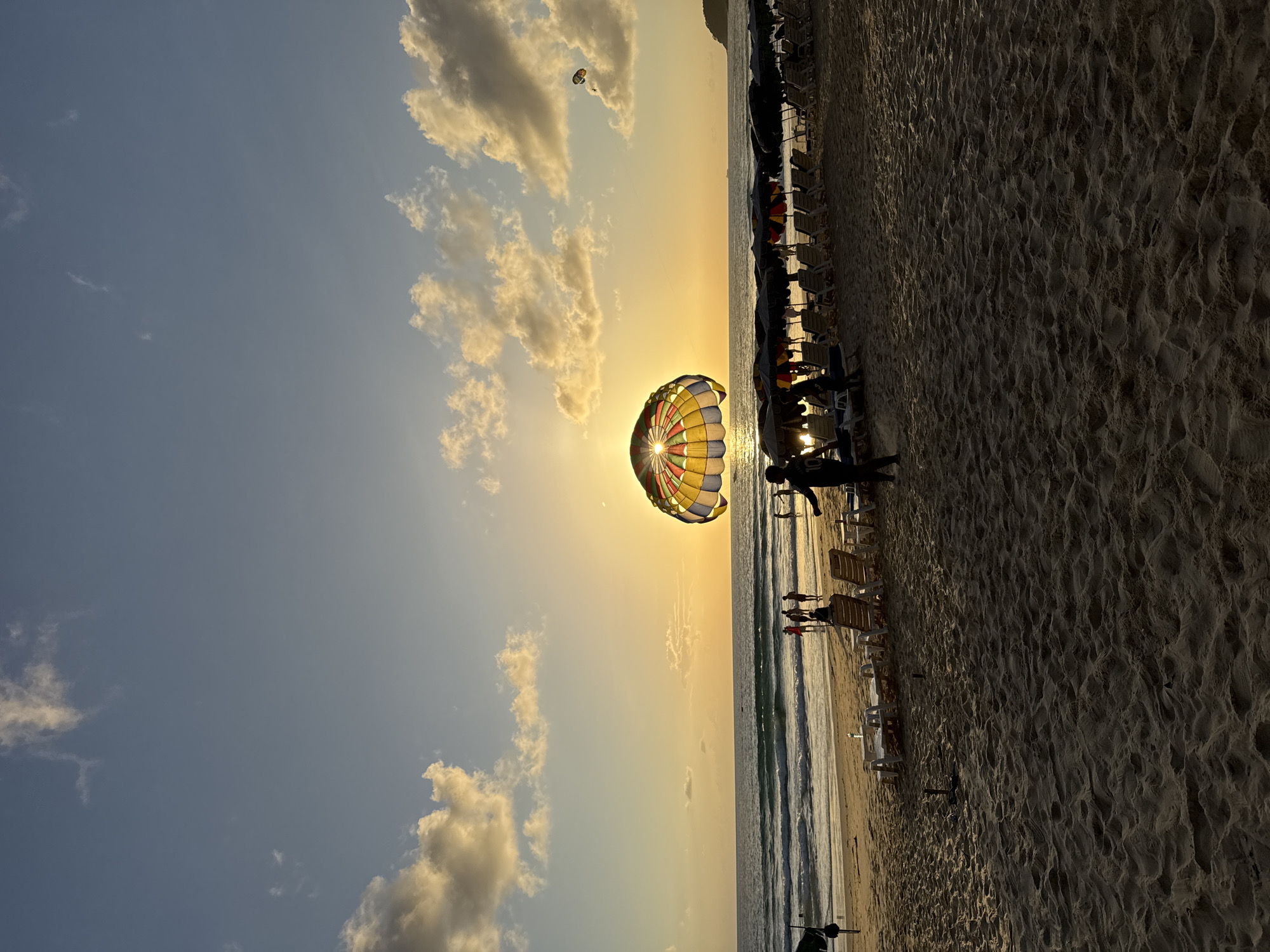 This image captures a sunset scene on Karon Beach in Karon, Thailand. The sun is low on the horizon, casting a warm golden glow across the sky and reflecting on the ocean waves. In the foreground, the sandy beach is dotted with lounge chairs and umbrellas, with several people walking or standing. A large, colorful parachute, likely used for parasailing, is prominent in the sky, with the sun partially obscured behind it. Another, smaller parachute is visible in the distance. The sea is calm with gentle waves rolling onto the shore. The overall atmosphere is serene and picturesque, indicative of a popular tourist destination during evening hours.