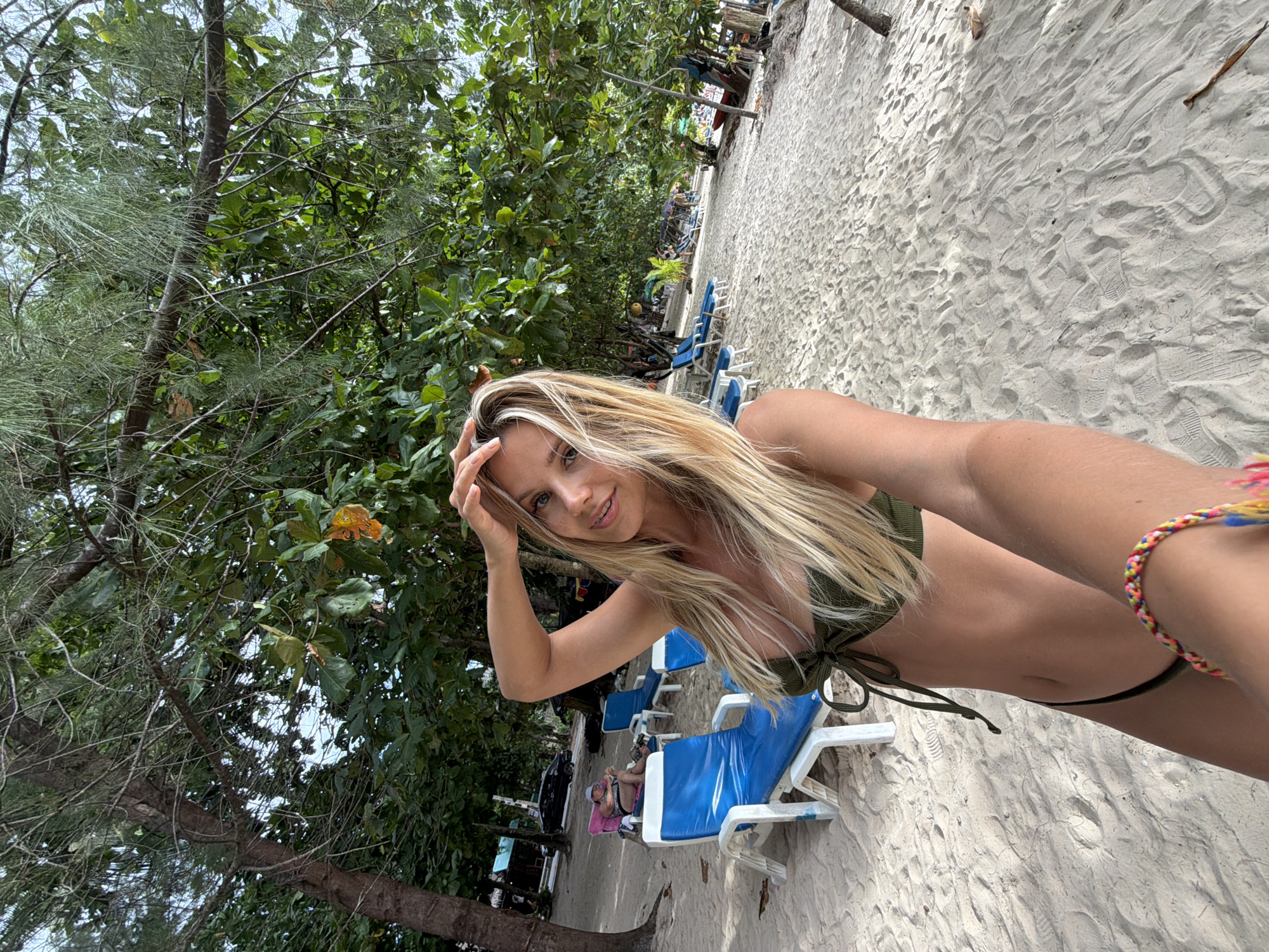 A blonde woman in a green bikini is taking a selfie on a sandy beach in Sakhu, Thailand. She is wearing a colorful bracelet on her left wrist and has her right hand touching her hair. In the background, there are rows of empty blue lounge chairs and trees with green foliage. The sky is overcast. A person is visible sitting in a pink lounge chair in the distance.