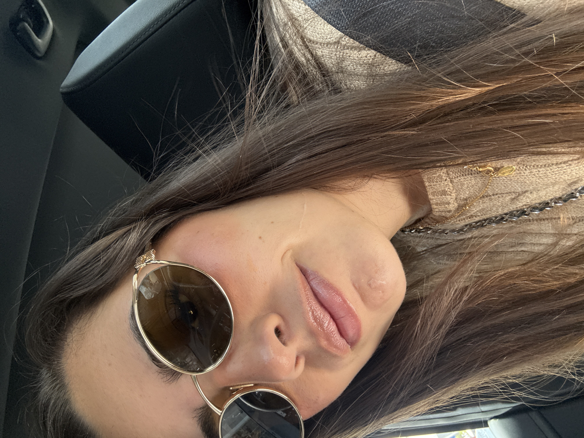 A woman with long brown hair and round sunglasses poses for a close-up selfie inside a vehicle. She is wearing a beige sweater with a chain detail. The vehicle's black interior is visible in the background. There is no information to suggest the location is in Heroica Puebla de Zaragoza, Mexico.