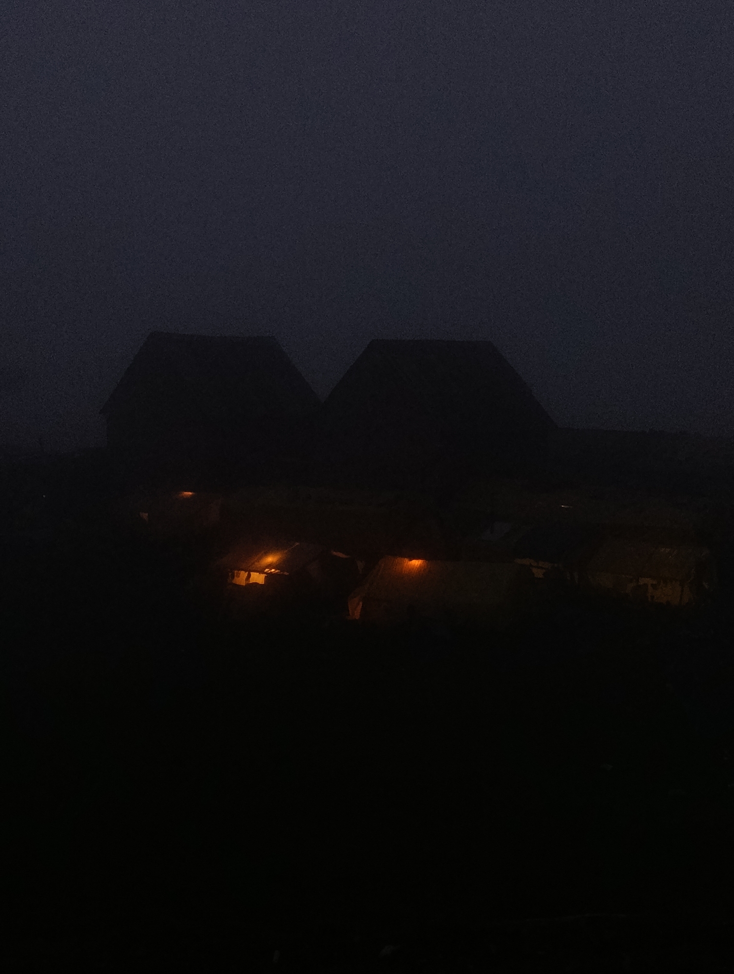 The image shows a dark, foggy scene with two dimly lit buildings in the foreground. The buildings appear to be traditional, with steep roofs. There are visible lights within the buildings, suggesting occupancy. The surrounding environment is obscured by darkness and fog, making it difficult to discern specific details of the location. The overall impression is one of a quiet, rural setting at night. While the image is labeled as being in Marhi, India, there are no specific visual cues in the photograph to confirm this location.