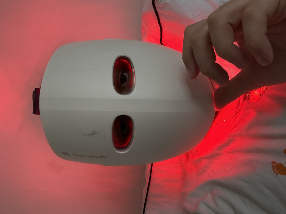 An individual is depicted indoors in the city of London, United Kingdom, wearing a white Therabody light therapy mask. The mask covers the entire face, featuring two horizontal oval apertures through which the wearer's eyes are visible. The brand name "Therabody" is printed vertically on the left side of the mask, which also exhibits minor smudges on its upper left surface. Intense red light emanates from the mask's interior and lower edge, casting a strong red hue on the immediate surroundings, including a plain background wall and white fabric beneath the mask. A human hand, positioned in the lower right quadrant of the image, reaches towards the bottom edge of the mask. A thin chain necklace with a small, reflective pendant is visible around the wearer's neck, partially obscured by the red light. The white fabric beneath the mask, possibly clothing or bedding, features a black trim and an orange footprint logo. Black wires are partially visible in the background, to the left and right of the mask's upper portion.