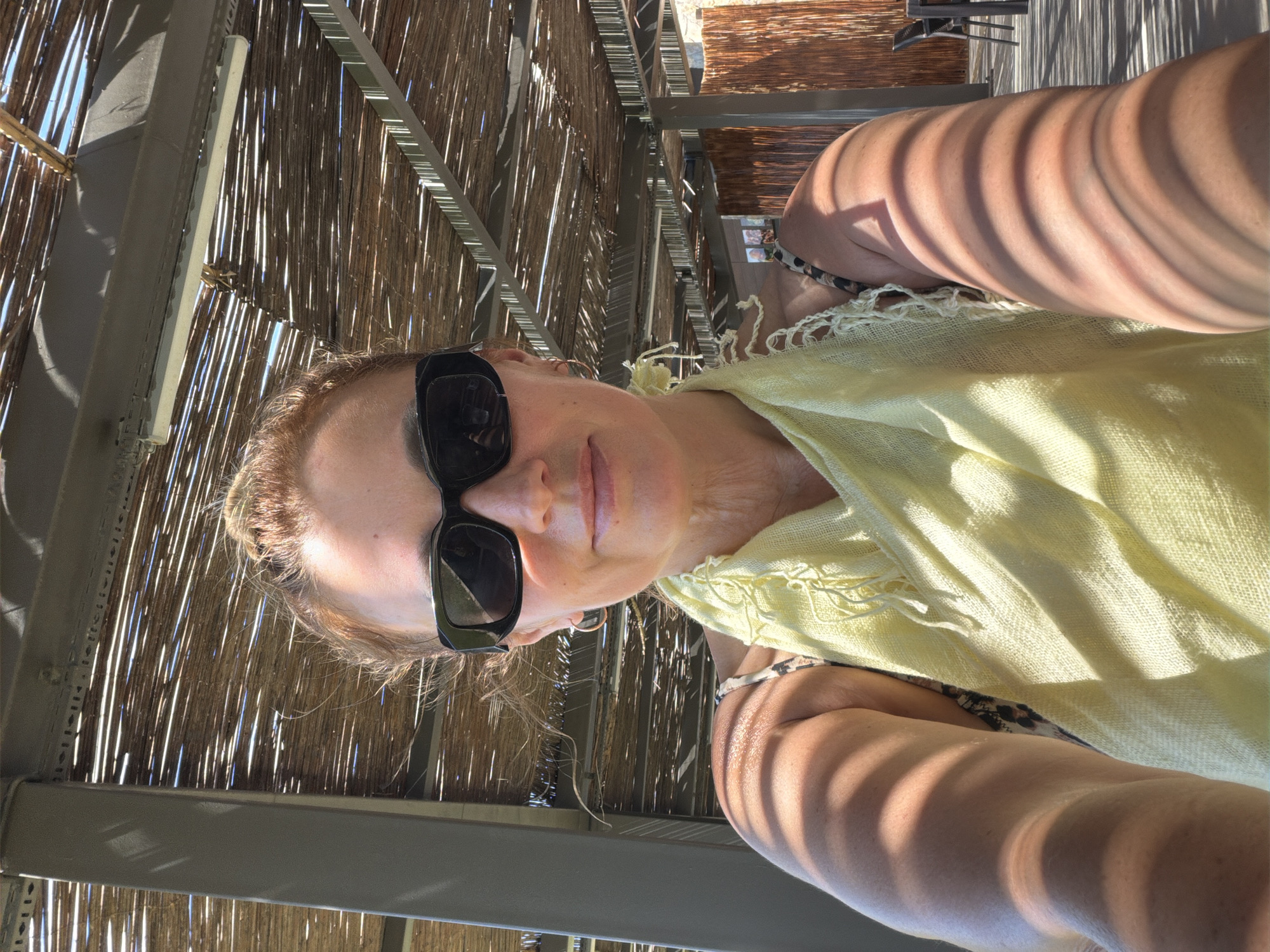 A woman with light-colored hair pulled back is wearing black sunglasses, a light yellow scarf, and a patterned top. She is looking into the camera and holding it out to take a selfie. She is outdoors under a covered patio with a thatched roof. The sun is casting shadows on her face and arm. In the background, there are wooden walls and outdoor furniture.