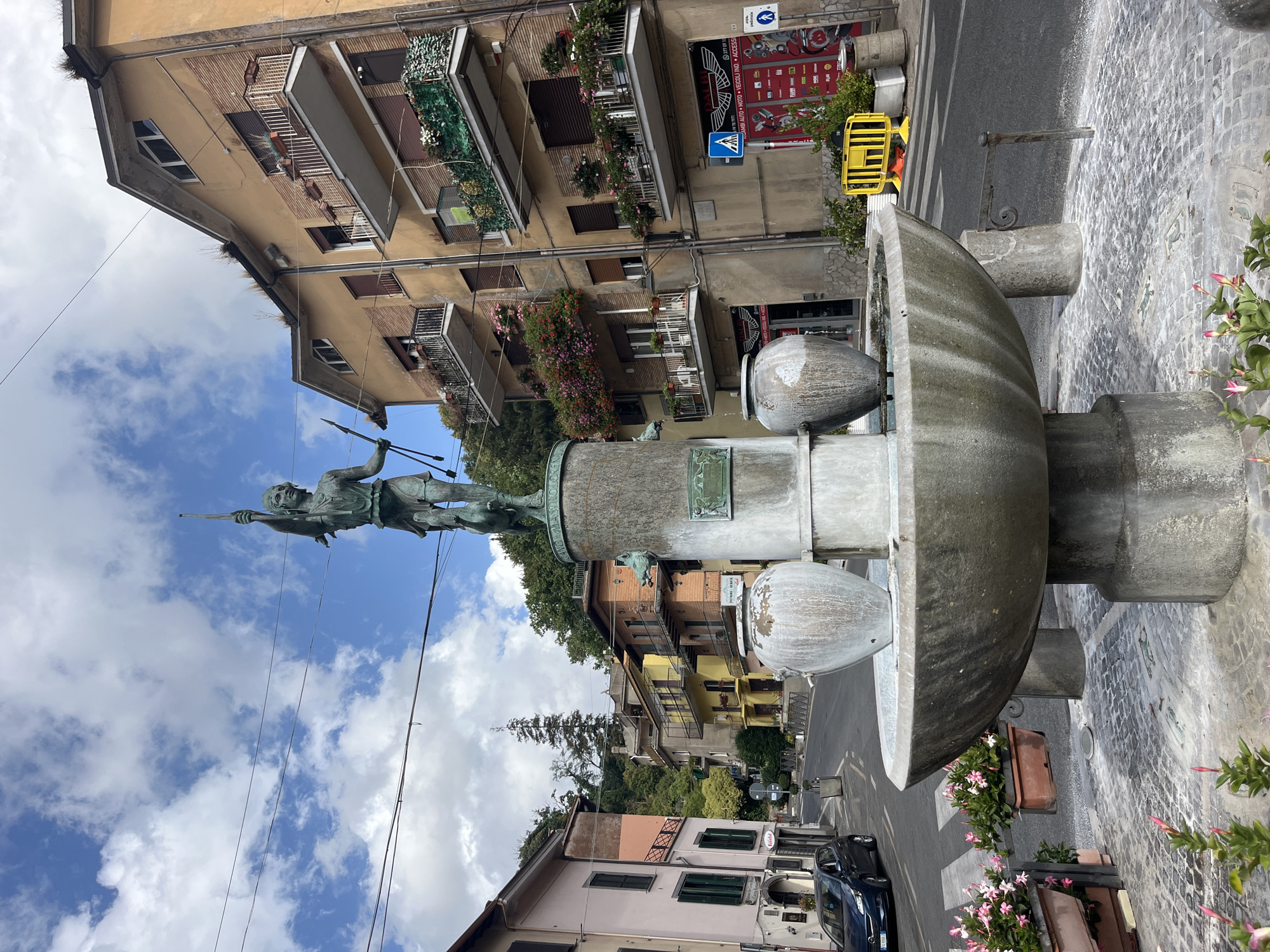 The image captures a daytime street scene in Nemi, Italy, featuring a prominent fountain in the foreground. The fountain is made of concrete and stone, with a statue of a figure holding spears atop a pedestal. Two large urns are positioned on the fountain's structure.
The fountain is situated on a cobblestone plaza, bordered by a street. Buildings line the street, displaying a variety of architectural styles and colors, including apartments with balconies adorned with flowers. A dark-colored car is parked on the left side of the street.
In the background, lush green trees and hills suggest a suburban or semi-rural setting. The sky is partly cloudy, with a bright blue expanse and scattered white clouds. The overall impression is of a charming, tranquil Italian town square.