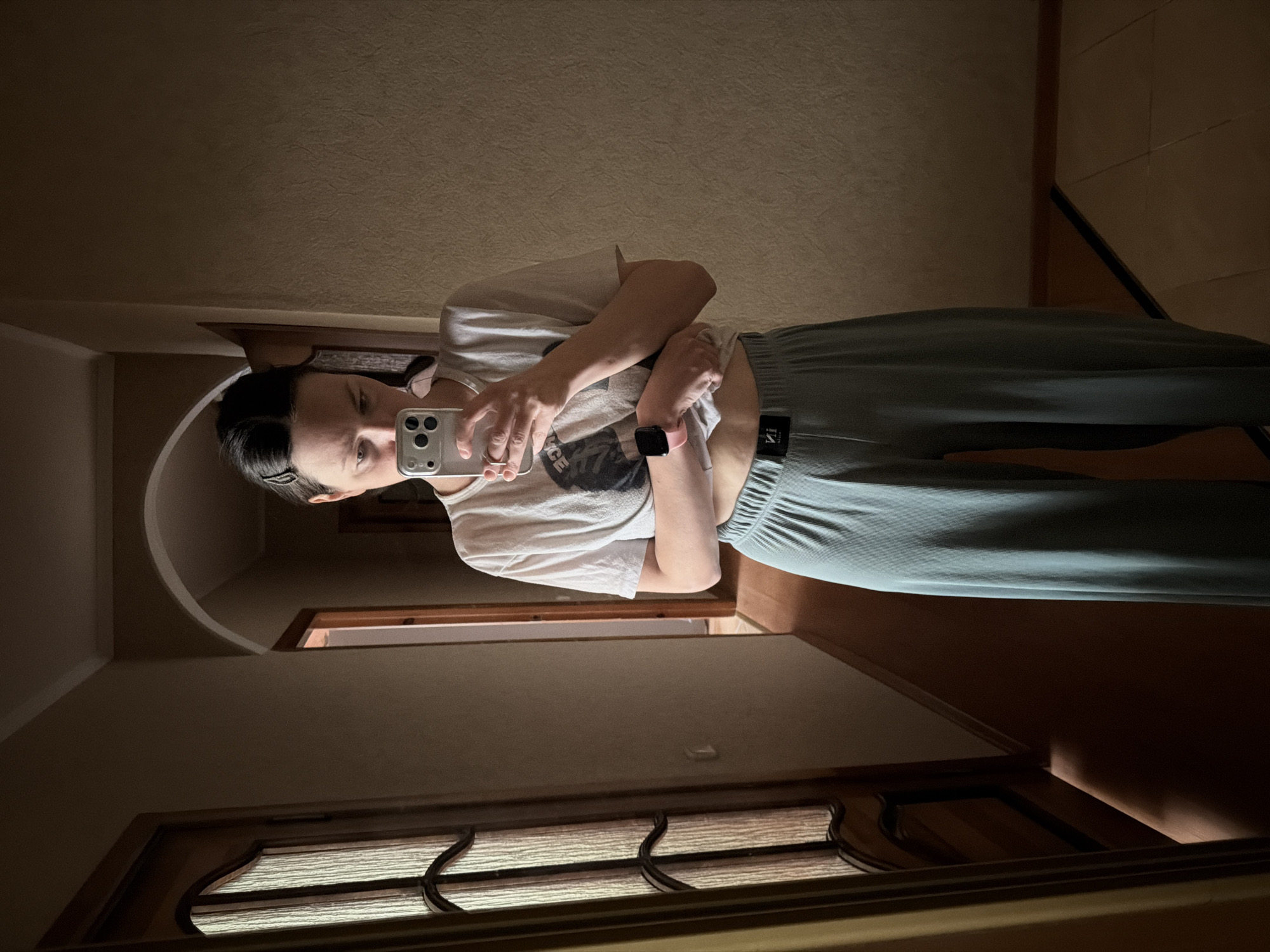 A woman is taking a mirror selfie in a dimly lit room of a city of Unknown, Unknown. She is holding a phone in her right hand, wearing a light-colored t-shirt and gray sweatpants. The room is dark, illuminated only by a soft light source. Details about the building or specific location are indiscernible.
