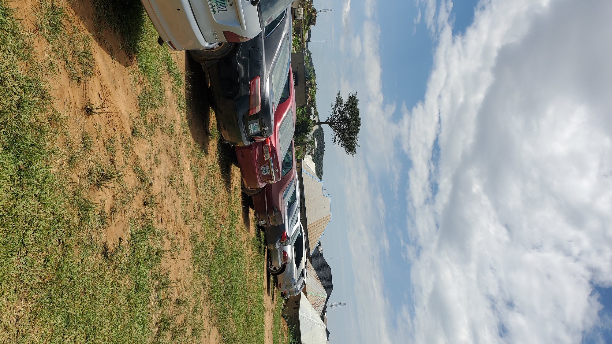 A vertical, eye-level outdoor shot shows a row of parked cars on an unpaved lot in Barakin Akawo, Nigeria. The cars, a mix of sedans, are parked nose-in, facing away from the camera towards some buildings in the background. The buildings, with corrugated metal roofs, are modest and appear to be residences or small commercial structures. A single, mature tree with a spreading canopy stands alone on a rocky outcrop behind the cars, silhouetted against a bright, cloudy sky. The ground is a mix of dry, reddish-brown earth and patches of green grass. The overall impression is of a sunny day in a rural or semi-rural area.
