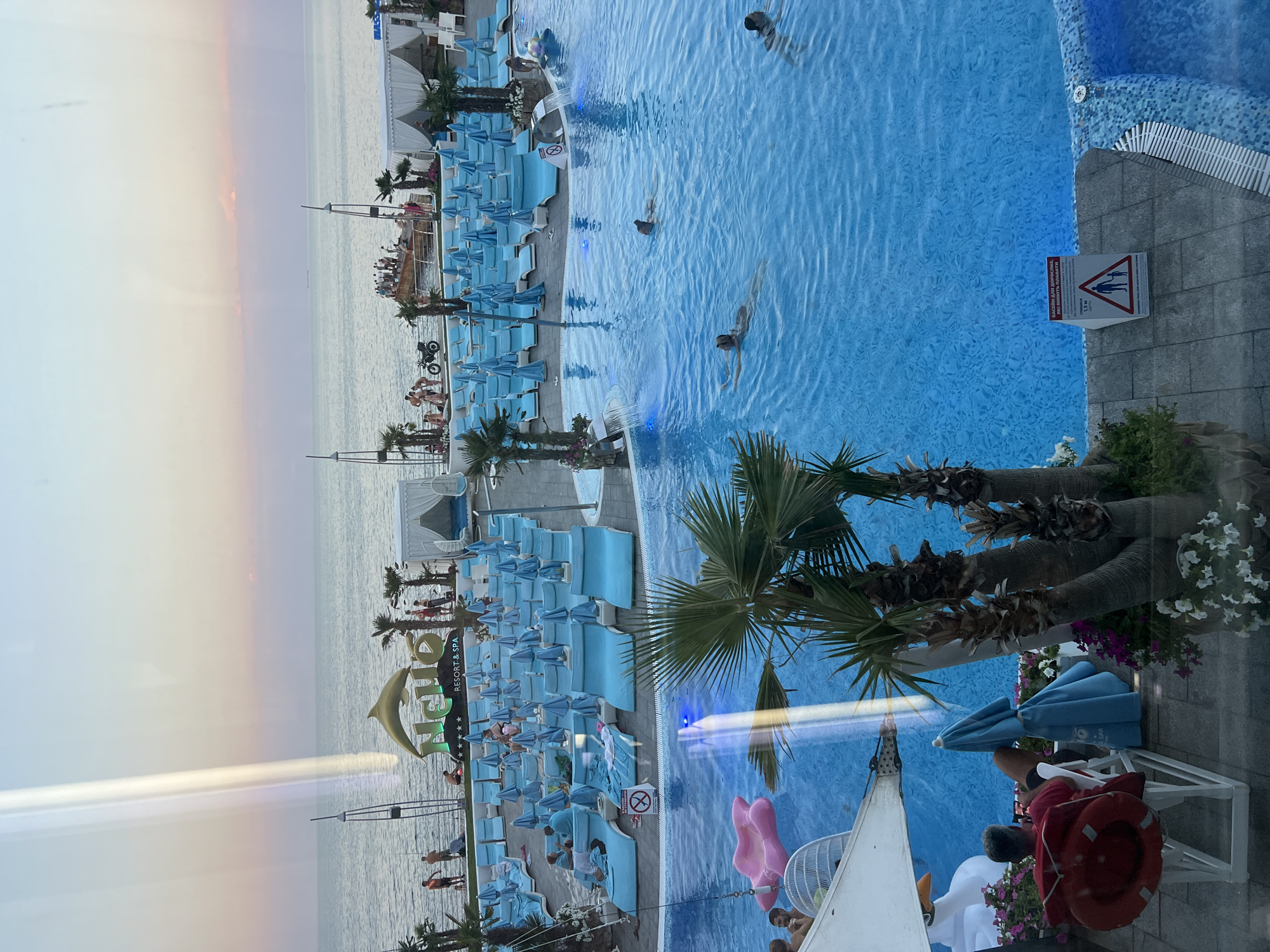 This image depicts a resort area in Odesa, Ukraine, overlooking the Black Sea. In the foreground, a large blue swimming pool is visible with several people swimming. To the left, a man in a red shirt sits in a lounge chair, holding a phone. In the middle ground, rows of blue lounge chairs and umbrellas are set up on a paved area, with palm trees interspersed. A sign for "NEMO RESORT & SPA" with a dolphin motif is prominently displayed. In the background, the Black Sea stretches to the horizon under a soft, pastel sky, with a few boats visible in the distance. The overall scene suggests a vacation or leisure setting.