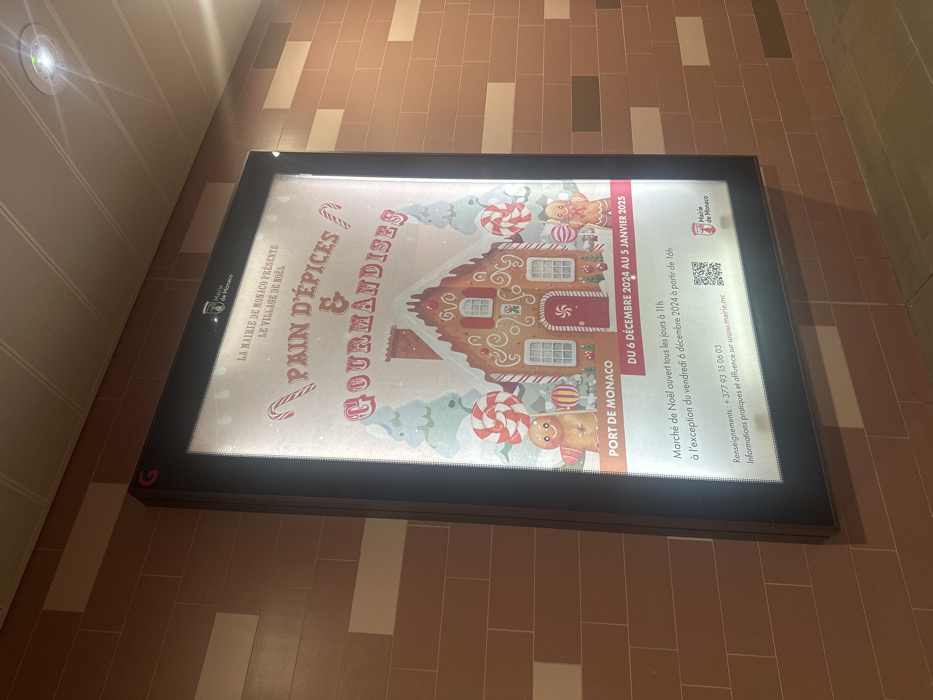 An illuminated sign lies flat on a tiled floor in what appears to be an indoor public space in Beausoleil, France. The floor consists of light brown and beige rectangular tiles arranged in a linear pattern. A bright overhead light fixture is visible on a paneled wall to the left, casting a glow.
The sign is a promotional poster for a Christmas event, featuring a festive illustration of a gingerbread house adorned with white frosting, candy canes, gingerbread figures, and other sweets against a light blue and white background.
Visible text on the sign indicates:
- "LA MAIRIE DE MONACO PRÉSENTE LE VILLAGE DE NOËL" (The Monaco City Hall Presents The Christmas Village)
- The main title reads "PAIN D'ÉPICES & GOURMANDISES" (Gingerbread & Delicacies).
- The location is specified as "PORT DE MONACO."
- The event dates are "DU 6 DÉCEMBRE 2024 AU 5 JANVIER 2025" (From December 6, 2024 to January 5, 2025).
- Details about the "Marché de Noël" (Christmas Market) state it is "ouvert tous les jours à 11h à l'exception du vendredi 6 décembre 2024 à partir de 16h" (open every day at 11 am, with the exception of Friday, December 6, 2024, starting at 4 pm).
- Contact information includes "Renseignements: +377 93 15 06 03" and a website for "Informations pratiques et affluence sur www.mairie.mc."
- The "Mairie de Monaco" logo is present in the top left and next to a QR code on the right.
The sign is brightly lit from within, contrasting with the slightly dimmer surroundings. No people are visible in the image.