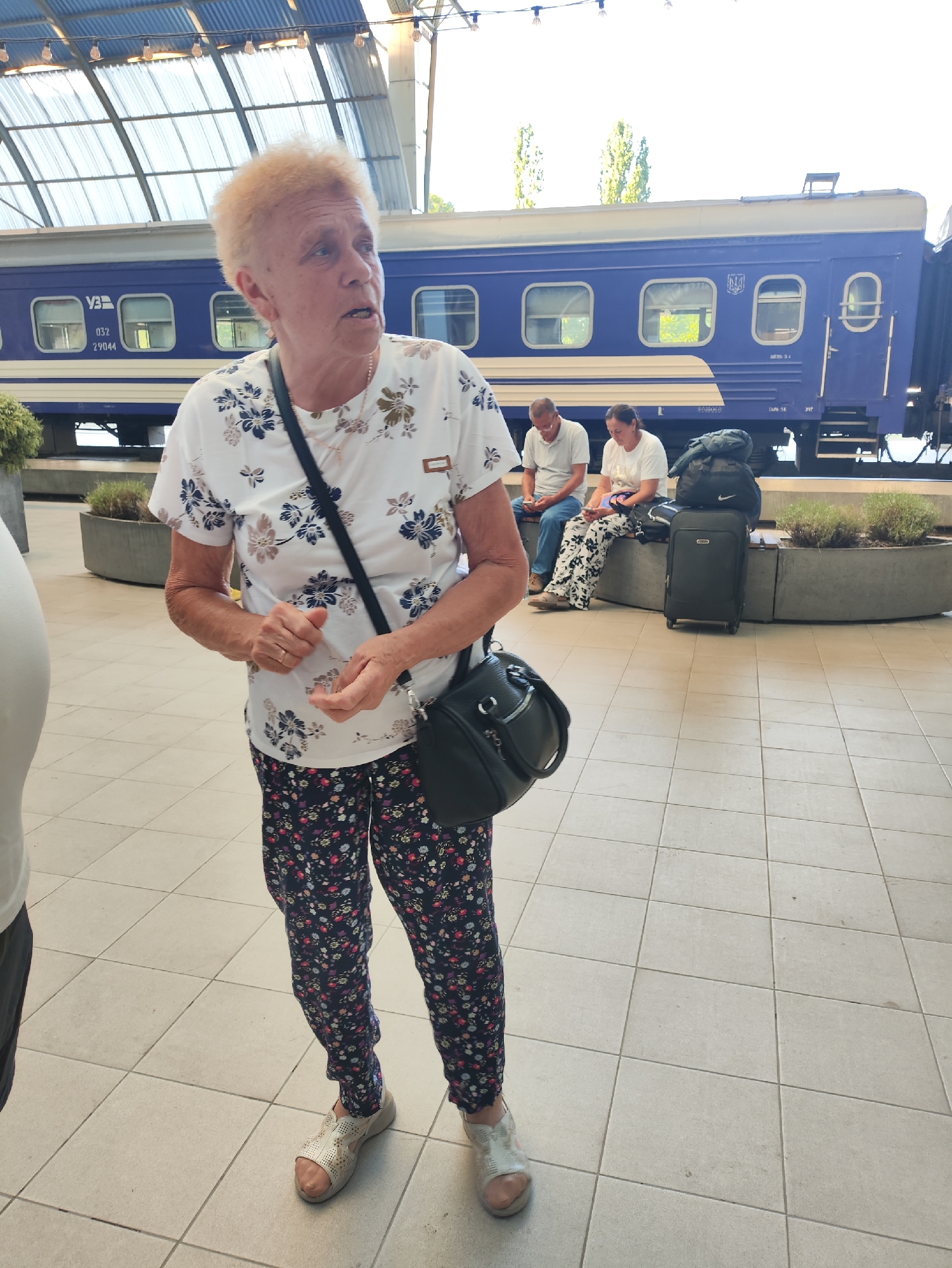 An older woman with short, curly blonde hair stands on a tiled platform at a train station. She is wearing a white t-shirt with a floral pattern of blue and brown flowers, and dark floral patterned pants. She has a black crossbody bag over her shoulder. Her hands are raised, and she appears to be speaking. Behind her, a blue train is visible, and two other people are sitting on a bench further down the platform. The environment appears to be an indoor or covered outdoor train station with a metal roof structure overhead. The scene is lit by natural daylight. There is no information in the image to suggest the location is the city of Chișinău, Moldova.