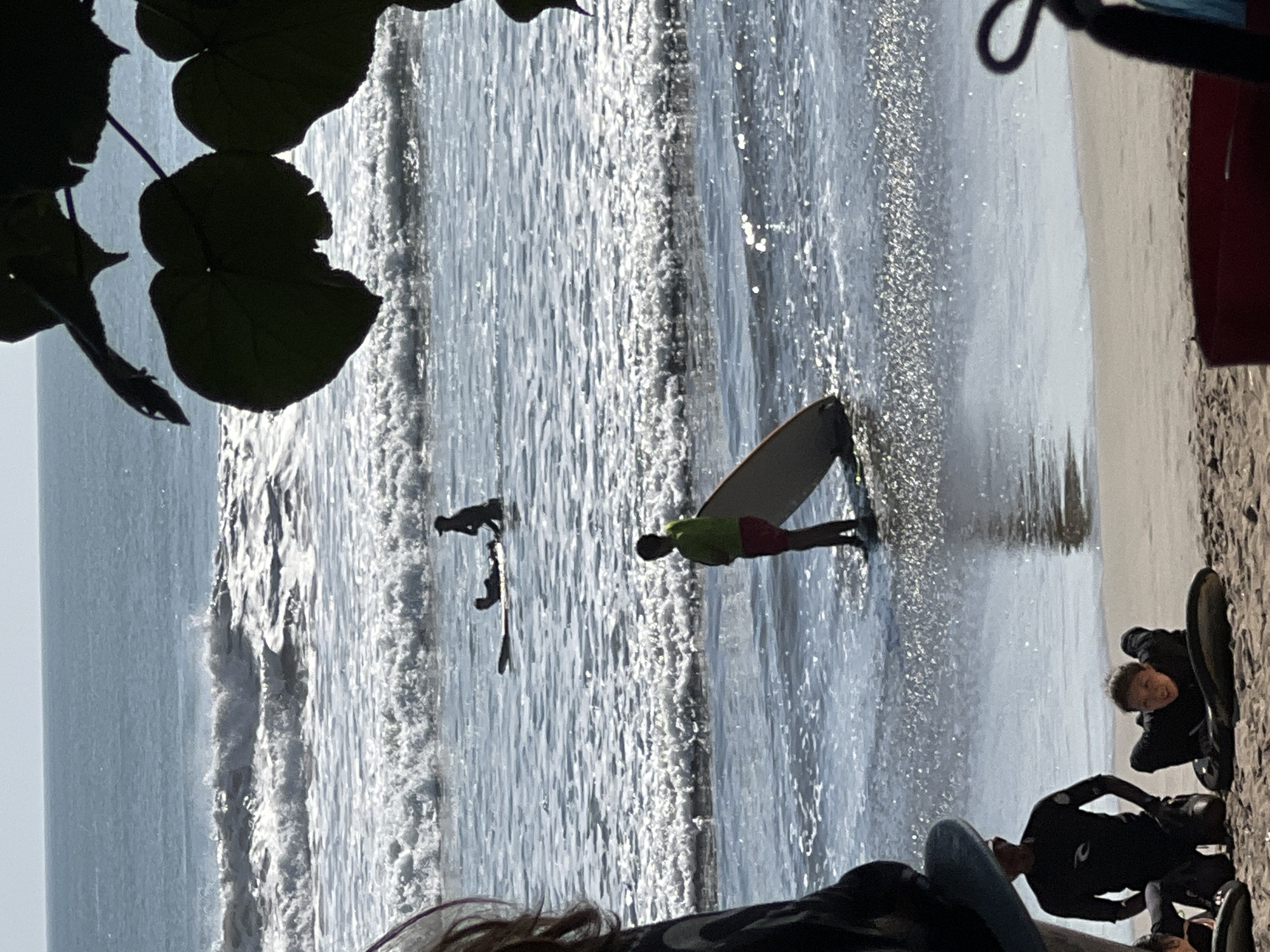 In Kuta, Indonesia, a sunny beach scene unfolds with surfers in the shallow water and on the sand. The ocean is moderately wavy, with white foam cresting on the incoming waves. In the water, three individuals are visible. One surfer, wearing a dark wetsuit, lies prone on a surfboard in the middle distance. Further out, another person, silhouetted against the bright sky, stands on a surfboard. Closer to the shore, a young person in a green shirt and red shorts stands with a surfboard, facing the ocean. On the sandy beach in the foreground, two individuals are present. One, wearing a black wetsuit, kneels on the sand. Another, also in dark attire, lies on a surfboard, facing forward. The scene is partially framed by dark green leaves in the upper right corner.