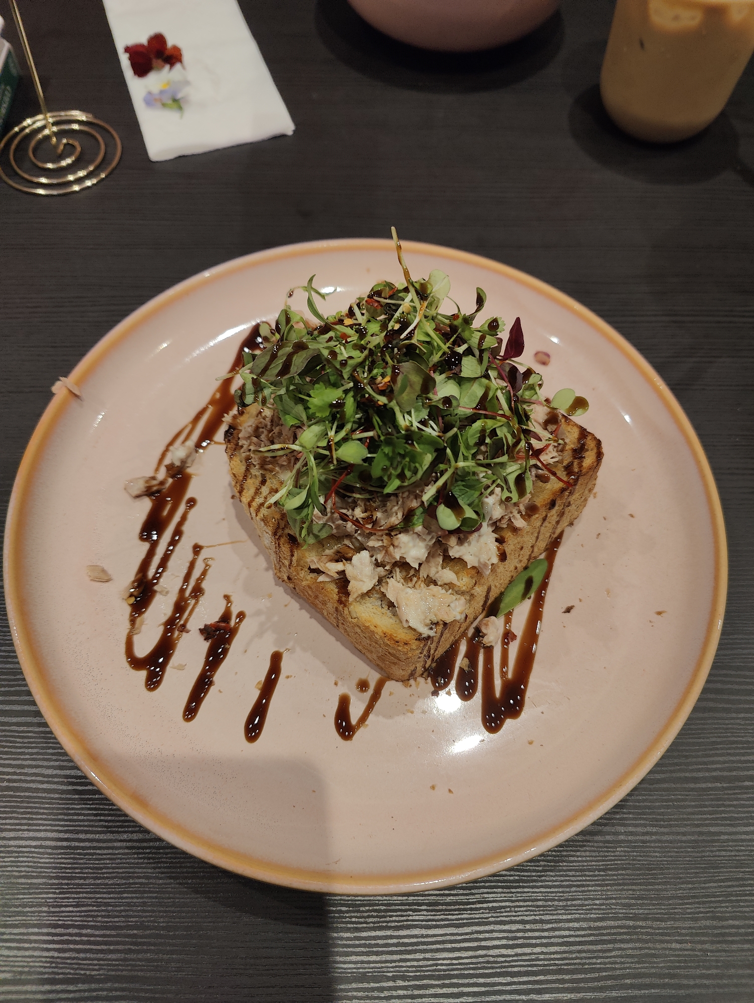 A high-angle shot shows a pale pink plate, edged in a darker pink, holding a piece of toast topped with a light-colored spread, possibly tuna or crab salad, and an ample heap of microgreens dressed with a dark sauce, possibly balsamic glaze. The sauce is drizzled artistically around the toast on the plate. In the background, a gold-colored spiral wire card holder is visible, along with a small white napkin holding a few red and white flowers. A beige drink in a glass or plastic container is also visible in the upper right of the image. The table surface is dark grey with horizontal lines. The image was most likely taken indoors at a restaurant or cafe, in Ipswich, United Kingdom. There are no people visible in the shot.