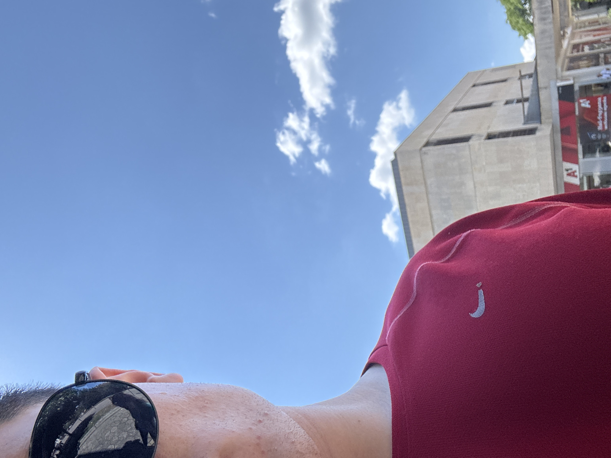 In the foreground of the image, a person's left side is visible, including their face, wearing dark sunglasses and a red t-shirt with a white Nike logo. The person's skin appears slightly sweaty. In the background, a concrete building with several windows is visible, and above it, a bright blue sky with scattered white clouds. The building appears to be part of a commercial area, with a sign that reads "A1" prominently displayed. This scene is located in the city of Kazanlak, Bulgaria.