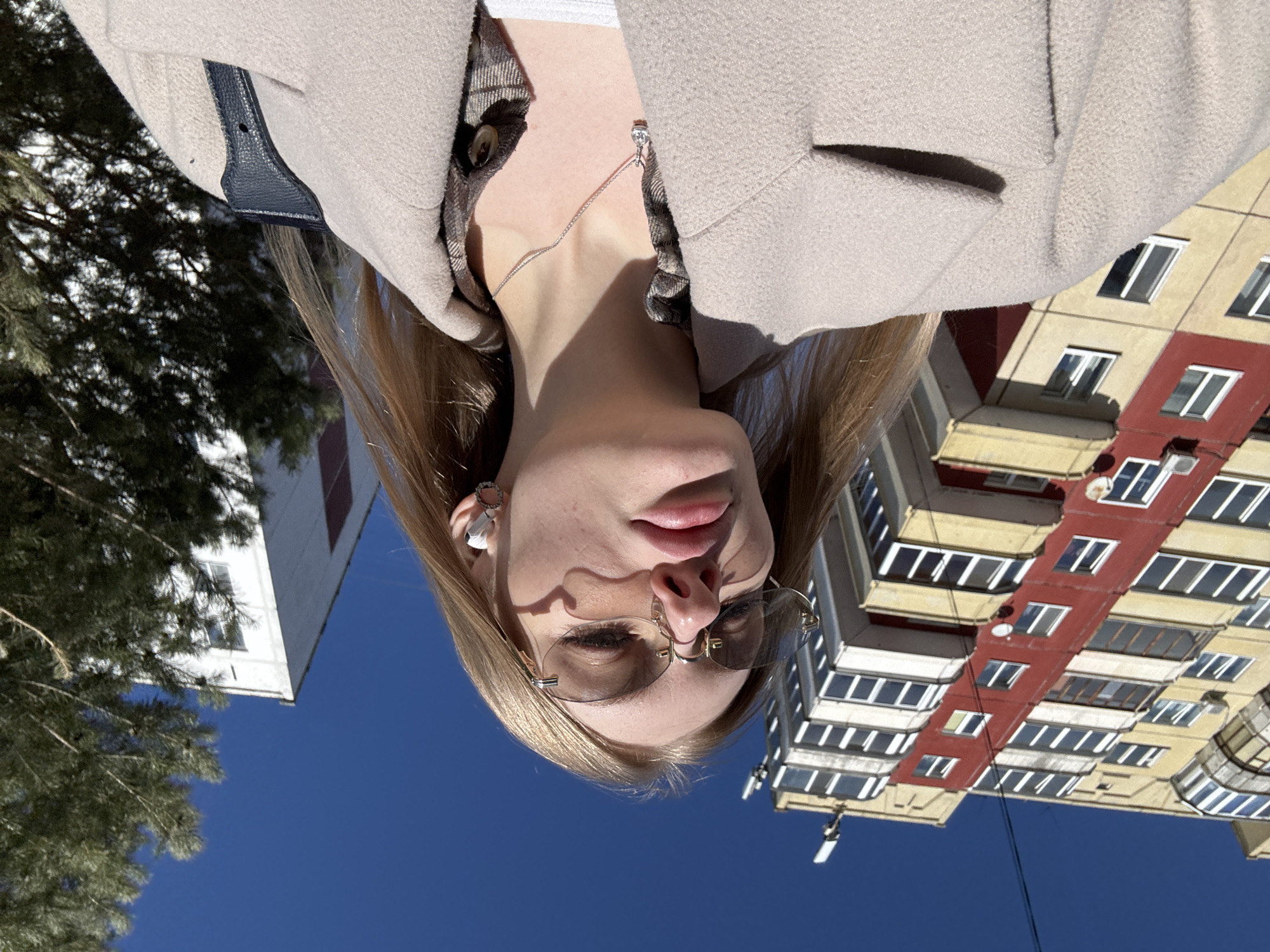 The image displays a low-angle outdoor portrait of a young woman situated in an urban environment in Perm, Russia, under a clear blue sky. The woman, positioned prominently in the foreground, has long, light-colored hair and is looking downwards. She is wearing rimless, light-tinted oval sunglasses with gold-colored frames. A white wireless earbud is present in her right ear, along with a small silver hoop earring. Her attire includes a light beige, collared wool-blend coat worn over a brown and beige plaid button-up shirt, with a white garment visible beneath. A silver chain necklace with a small, clear gemstone pendant is visible on her neck. A black strap, consistent with a shoulder bag or purse, rests on her right shoulder.
The background features two buildings and vegetation. To the left, a multi-story residential building dominates the frame, characterized by a yellow-beige facade with a distinct vertical red section. It features numerous windows and balconies, with communication antennas visible on its roof and an air conditioning unit on one balcony. To the right, partially obscured by a dark green evergreen tree, is a white, multi-story building.