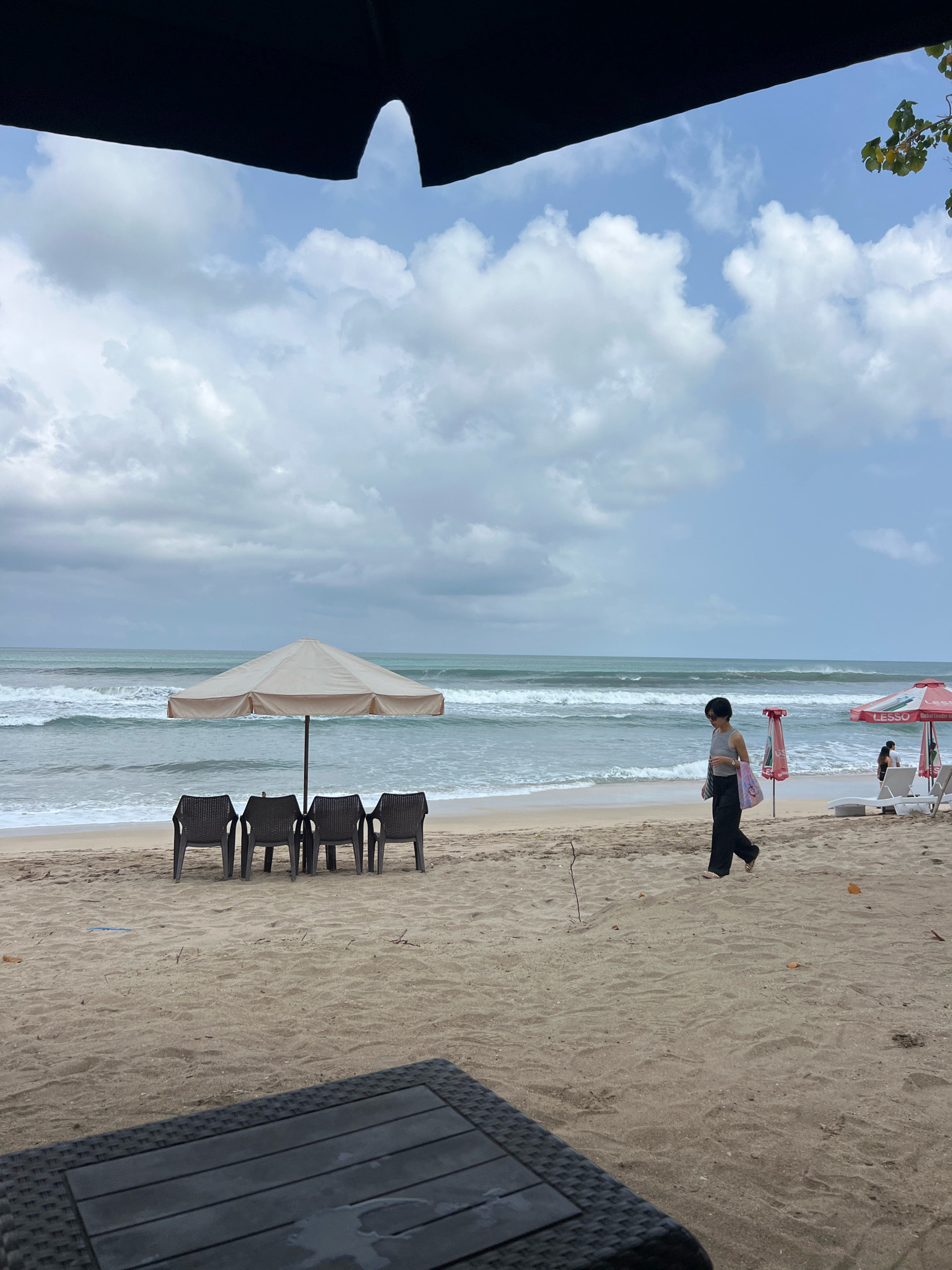 A woman wearing a grey tank top and black pants walks across a sandy beach in Kuta, Indonesia. She is carrying a patterned tote bag. In the background, the ocean has moderate waves, and the sky is cloudy. There is a beach umbrella with four chairs underneath it, and further down the beach, two red umbrellas and some lounge chairs are visible. The foreground shows the edge of a dark-colored outdoor table.
