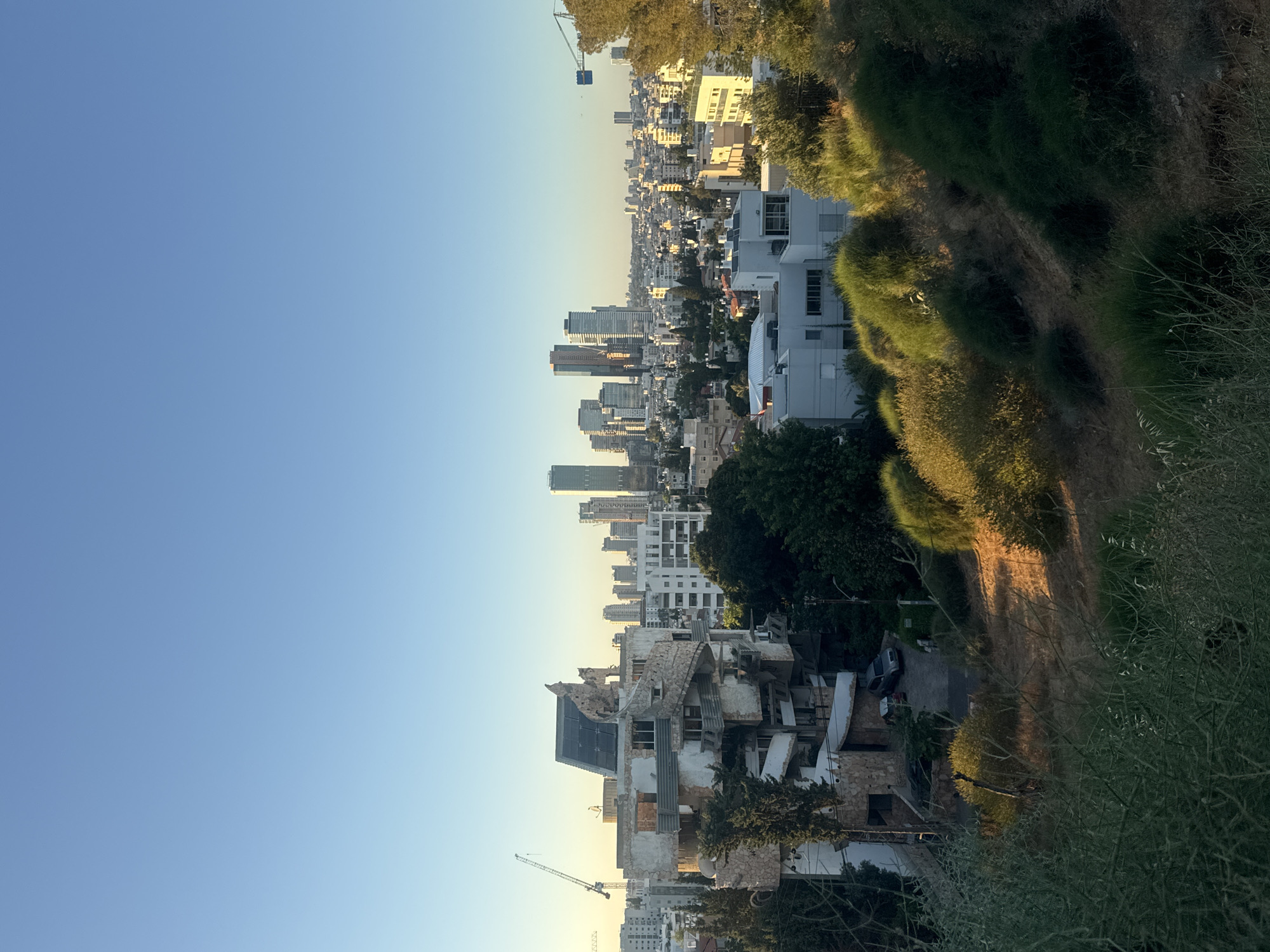 This image depicts a panoramic view of Ramat Gan, Israel, with a focus on a distinctive, multi-story building in the foreground. The building appears to be undergoing renovation or is in a state of disrepair, with exposed concrete, a stone spiral staircase wrapping around its exterior, and what seems to be solar panels on its roof.
In the midground, a dense urban landscape unfolds, characterized by a collection of mid-rise and high-rise buildings, indicative of a city center. A prominent cluster of modern skyscrapers dominates the skyline in the distance, suggesting a financial or business district. Construction cranes are visible, signifying ongoing development within the city.
The foreground and immediate midground are populated by lush green trees and dense shrubbery, with some areas showing dry, sun-baked earth. The vegetation creates a natural buffer between the viewer and the urban environment. The scene is illuminated by natural sunlight, casting long shadows and highlighting the textures of the buildings and the terrain, suggesting it is either early morning or late afternoon. The sky above is clear and blue, transitioning to a lighter hue near the horizon. No people are visible in the frame.