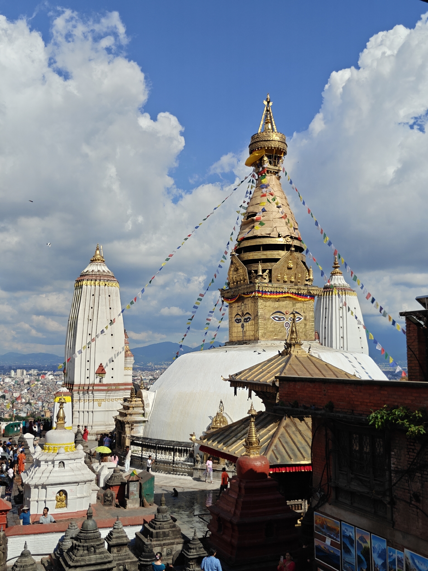 The image captures a wide view of the Swayambhunath Stupa complex in Kathmandu Metropolitan City, Nepal. The iconic stupa, with its golden spire and Buddha eyes, is prominent in the center, adorned with colorful prayer flags. To its left stands a tall, white stupa, and behind it, another white structure with a spire. In the foreground, smaller stupas and religious structures are visible, as well as numerous people walking around the complex. The city of Kathmandu sprawls in the background, with mountains visible on the horizon under a partly cloudy sky. The scene is bathed in daylight, with shadows indicating the time of day.