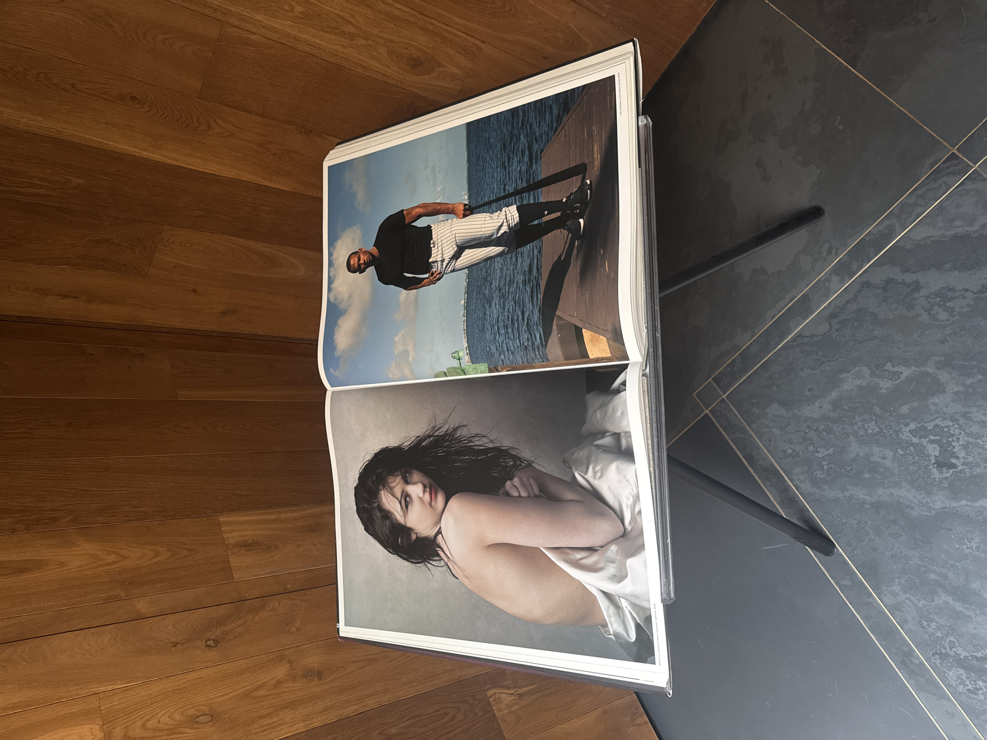 An open book rests on a black table, angled slightly off center. The book displays two separate photographic images. The top image features a Black man in a black shirt and vertically striped pants, holding a thin, long object. He stands on a wooden pier with water and sky in the background. The bottom image depicts a pale-skinned woman with dark, wet hair, partially covered by a white sheet.
The table sits on a tiled floor, with the tiles a dark gray with gold lines. To the left of the table is a wooden floor with light brown planks. The scene appears to be indoors. There is no indication this image was taken in Mexico City.