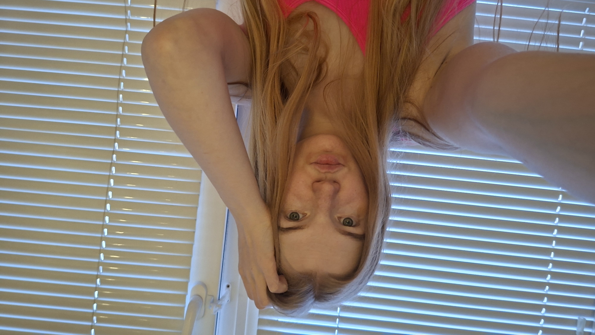 A woman with long blonde hair, wearing a pink top, is positioned upside down in front of a window covered with horizontal blinds. Her right hand is touching her forehead. The blinds suggest an indoor environment, likely a residential building. It is not possible to verify the location of the image in the city of Chaikovsky, Russia, from the visual information provided.