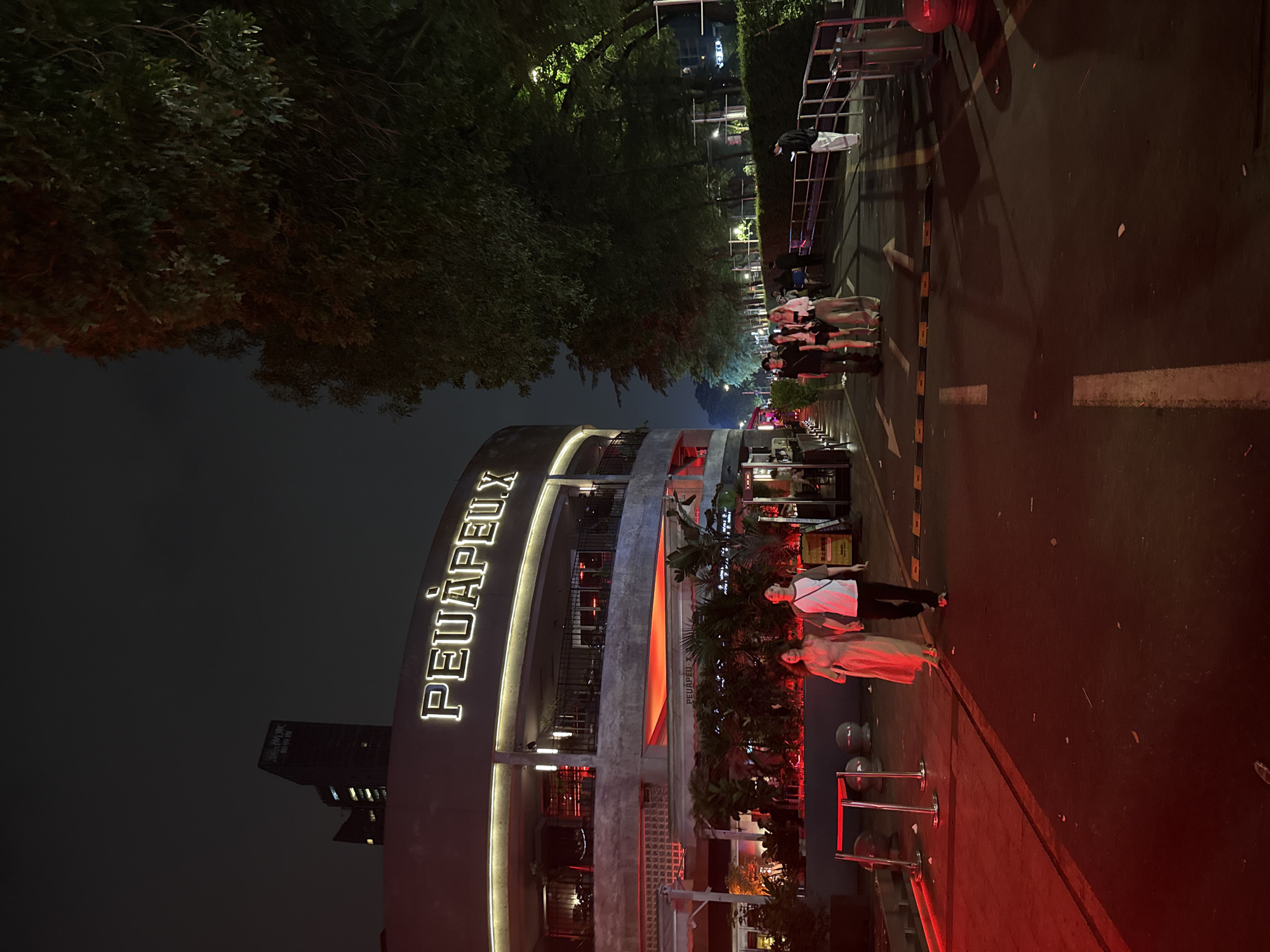 The image shows a street scene at night in Tianhe District, China, with the "PEUAPEUX" building prominently lit. The building is multi-storied and appears to be a commercial establishment, potentially a restaurant or bar. A group of people is walking along the sidewalk adjacent to the building, while another, smaller group is standing near the building's entrance. The street is paved and marked with lines. Lush greenery and trees line the upper left portion of the image. A portion of a taller building is visible in the background. The overall scene is well-lit by streetlights and building lights.