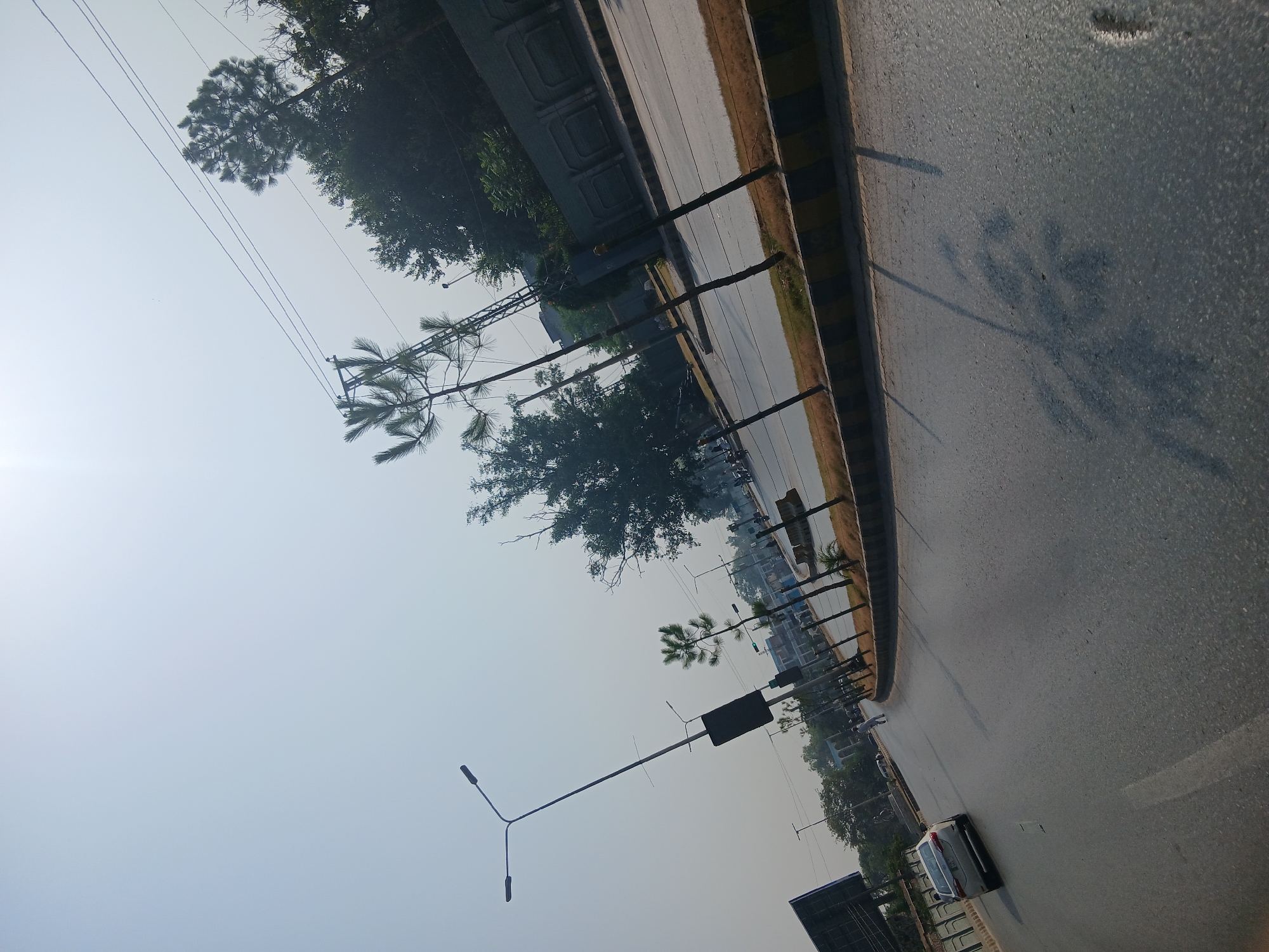 The image depicts a street scene in Peshawar City Tehsil, Pakistan, under a hazy sky. The perspective is from a moving vehicle, as indicated by the road surface filling the lower portion of the frame and the blurred shadows of trees and a lamppost cast upon it. On the left side of the road, a white sedan is visible, traveling in the same direction as the camera. Further down the road, a person is walking. The road is lined with trees and lampposts, and a concrete barrier with yellow and black markings separates the road from a grassy median. In the background, buildings and more trees can be seen. Power lines crisscross the sky. The overall impression is that of a typical urban street on a bright, possibly warm, day.