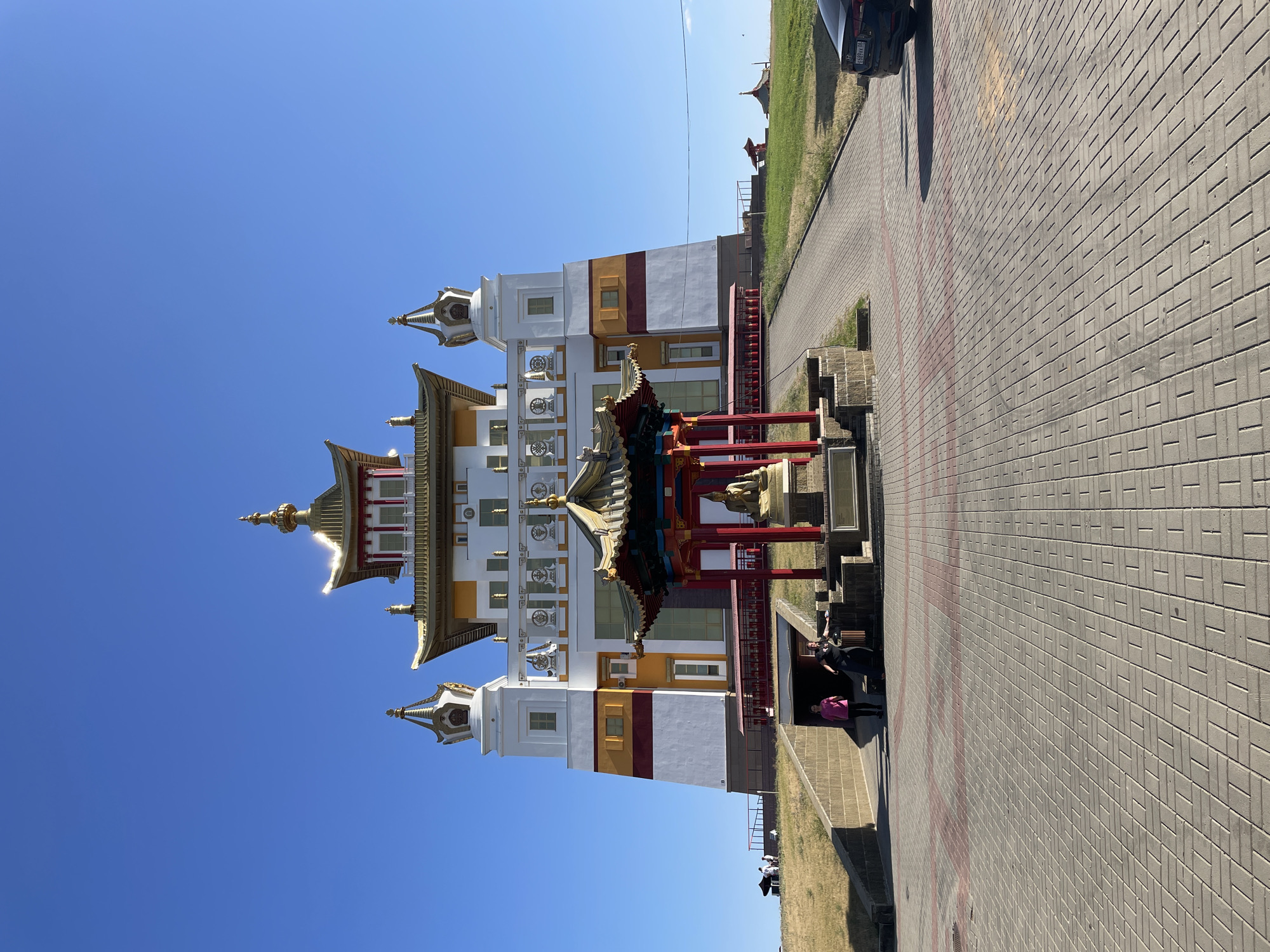 The image captures the Golden Temple of Elista, Russia, a prominent Buddhist structure. The temple is a multi-tiered building with a white base, adorned with yellow and red horizontal stripes on its lower sections. Its roofs are prominently gilded, featuring intricate Asian architectural elements. In front of the temple, there's a smaller, ornate pavilion housing a golden statue of Buddha.
The foreground is paved with grey interlocking bricks, with some red brick accents forming diagonal patterns. A paved ramp leads up towards the temple's entrance area. On the left side of the ramp, a group of three people are visible, standing and interacting. Two women are talking, while a man stands slightly apart. Further up the ramp, two women are walking up, one in a pink shirt and the other in dark clothing.
The temple is situated on a grassy incline. In the background, a clear blue sky dominates the scene. A dark-colored sedan is parked on the right side of the paved area, its rear visible. The overall impression is of a sunny day at a significant cultural and religious landmark.