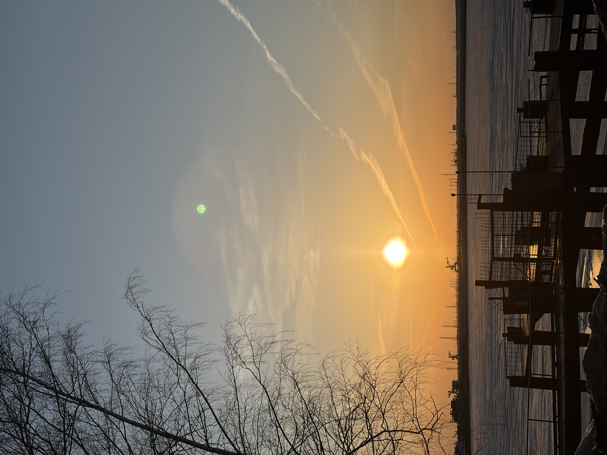 The image depicts a sunset scene in Chaikovsky, Russia. A bright sun is positioned over a body of water, possibly a frozen river or lake, with a warm orange glow in the sky. There are contrails visible in the sky. The foreground includes bare trees silhouetted against the sky. The right side of the image shows dark structural elements, possibly parts of a bridge or industrial facility, with a backdrop of industrial objects like cranes and electrical towers along the horizon.