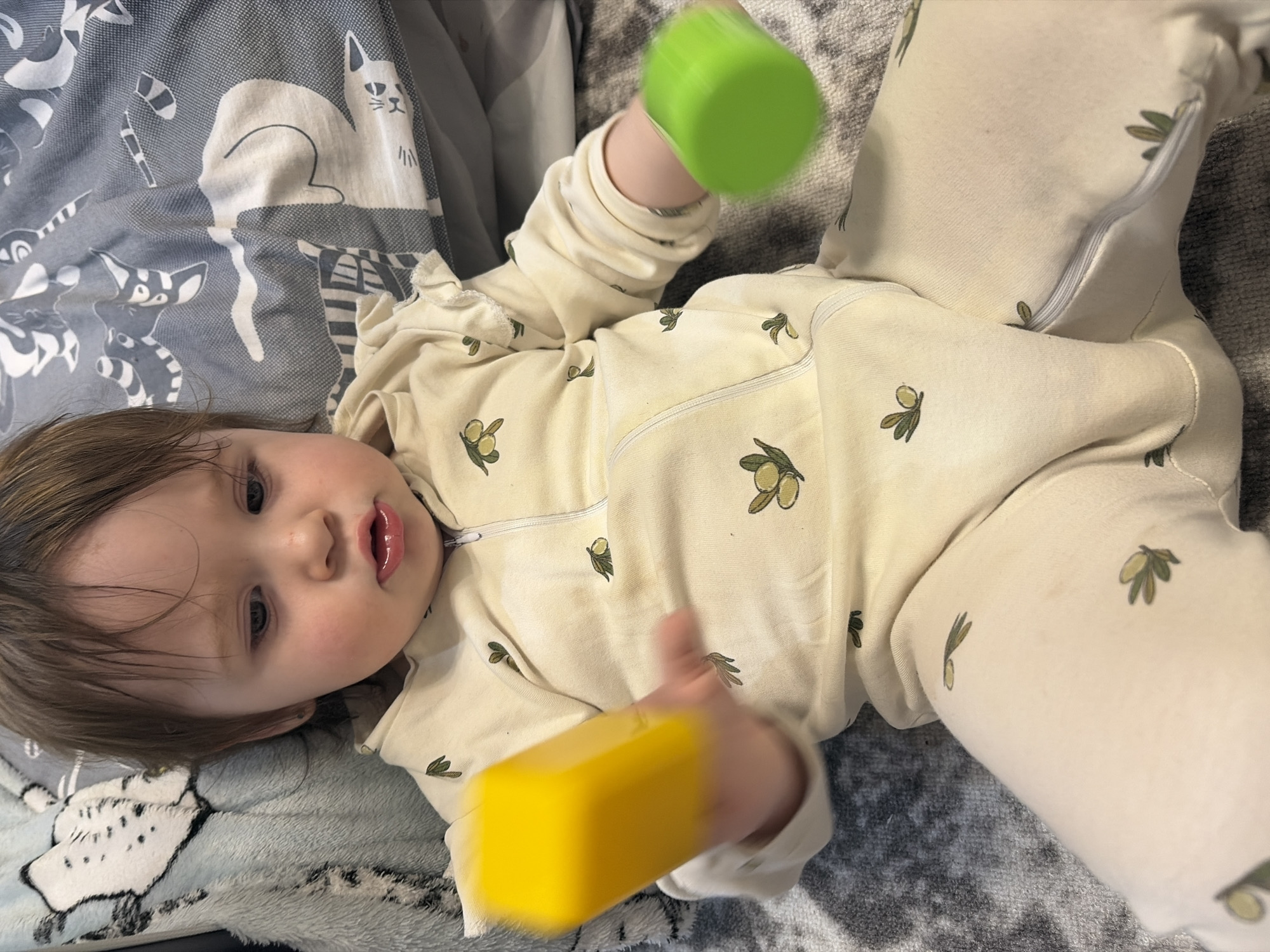 A baby lies on a patterned blanket, playing with yellow and green toys. The baby wears a white onesie with an olive branch pattern. The blanket features a pattern of cats. The location is not discernible from the image.
