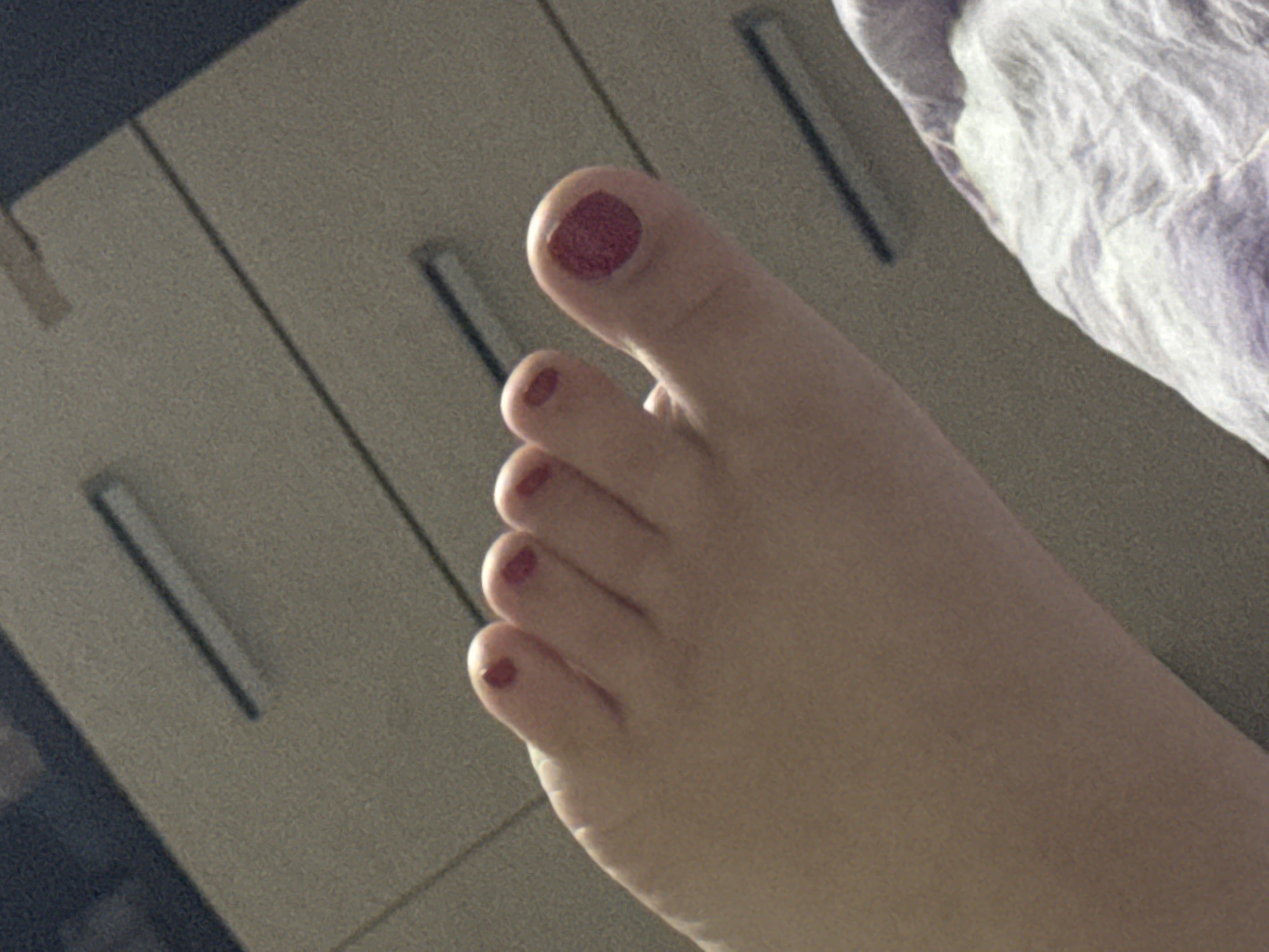The image shows a close-up of a human foot with toenails painted a dark red color. The foot is positioned on a light-colored surface, possibly fabric, with a textured background behind it. The background appears to be a wall with horizontal lines, suggesting a room or an interior space. The lighting is somewhat dim, casting soft shadows. There is no indication of the location being Saratov, Russia.