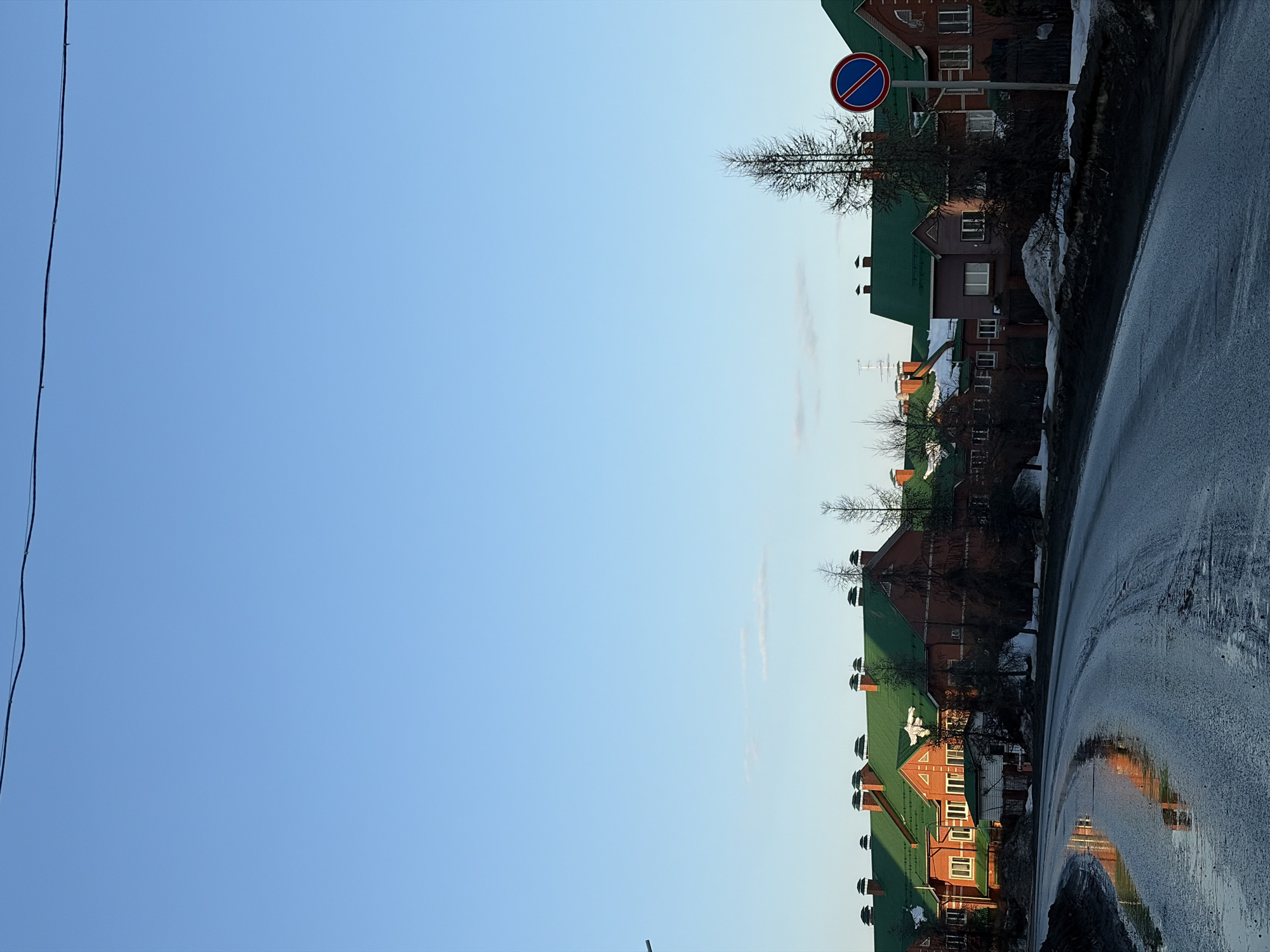 The image shows a street scene in Chaikovsky, Russia. On the right side of the frame, a wet asphalt road curves into the distance, reflecting the houses across the street. A row of red brick buildings with green roofs lines the road. A blue and red "No Stopping" traffic sign is visible. A power line crosses the upper left of the frame against a clear blue sky. Patches of snow remain along the roadside.