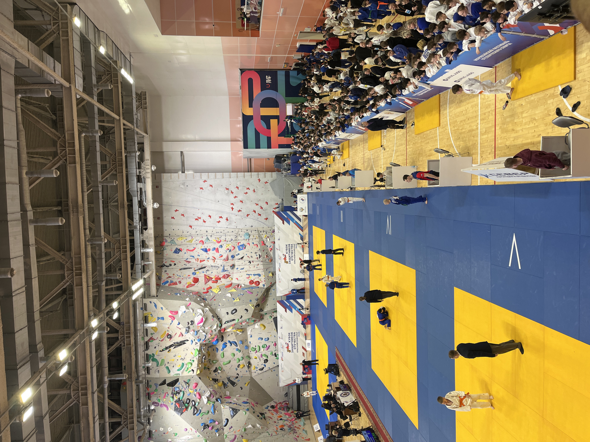 This image depicts a judo competition in Krasnoyarsk, Russia, as indicated by the signage. The judo mats are blue and yellow, with multiple matches potentially occurring simultaneously. A crowd of spectators, mostly young people in judo uniforms, is seated along the perimeter of the mat area.
In the background, there is a large climbing wall, which suggests the event is taking place in a multi-purpose sports facility. A banner with the text "ВСЕРОССИЙНСКАТА Фестиваль Студенческого СПОРТА DORUS JUDO" (All-Russian Festival of Student Sports) is visible near the spectators. Several officials or coaches, dressed in suits or uniforms, are present on the mat area. The structural support beams of the building are exposed on the left side of the image, indicative of a large indoor venue.