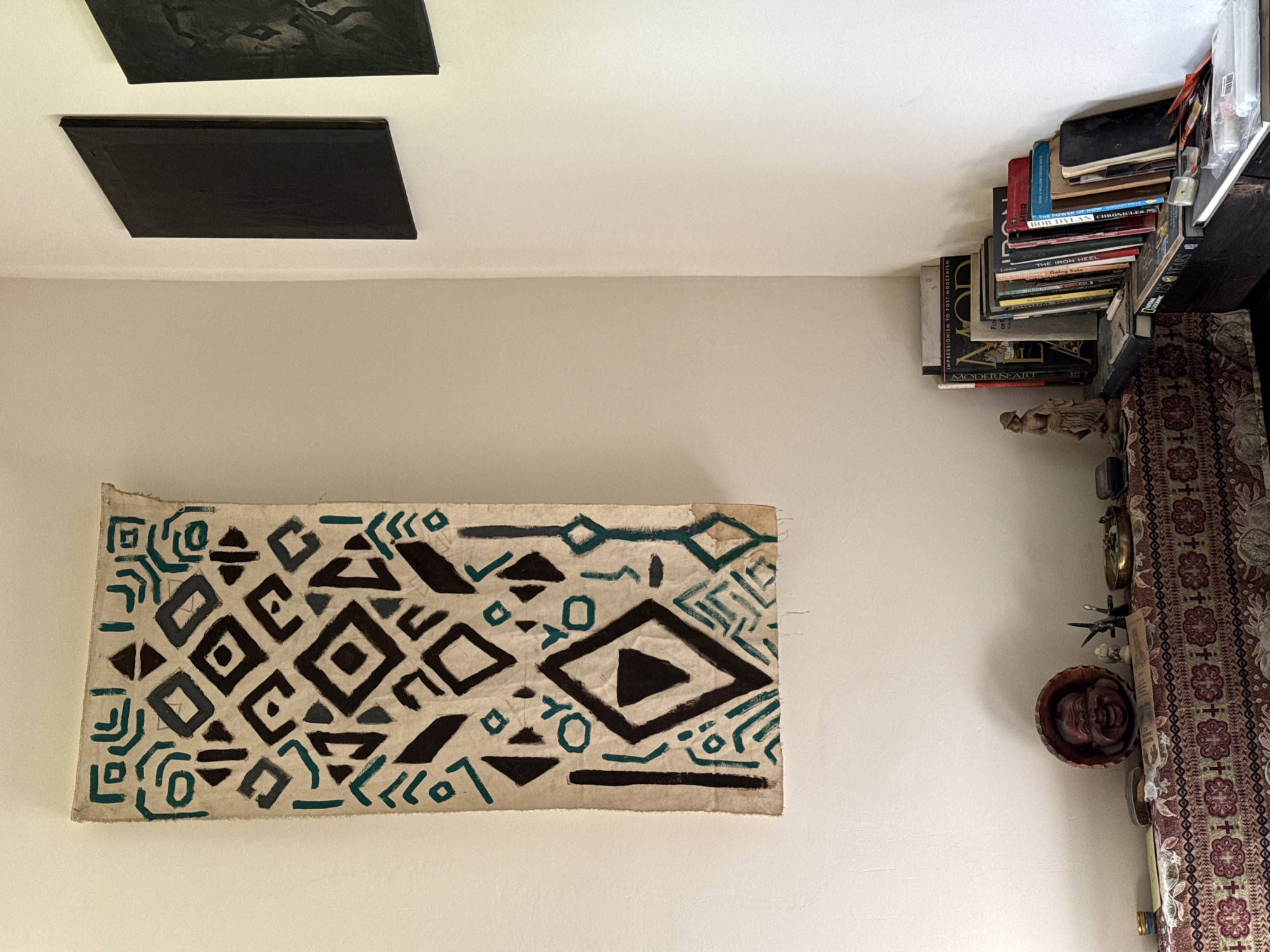The image presents an interior space. Two black square canvases hang on the white ceiling in the upper left corner. On the off-white wall below, there is a rectangular textile with a black and teal geometric pattern. Adjacent to this wall, stacked books sit atop a partially visible shelf, which is adorned with decorative items like a carved wooden face in a bowl and a patterned runner. The scene takes place indoors. There is no indication of a specific location in Karaberd, Armenia.