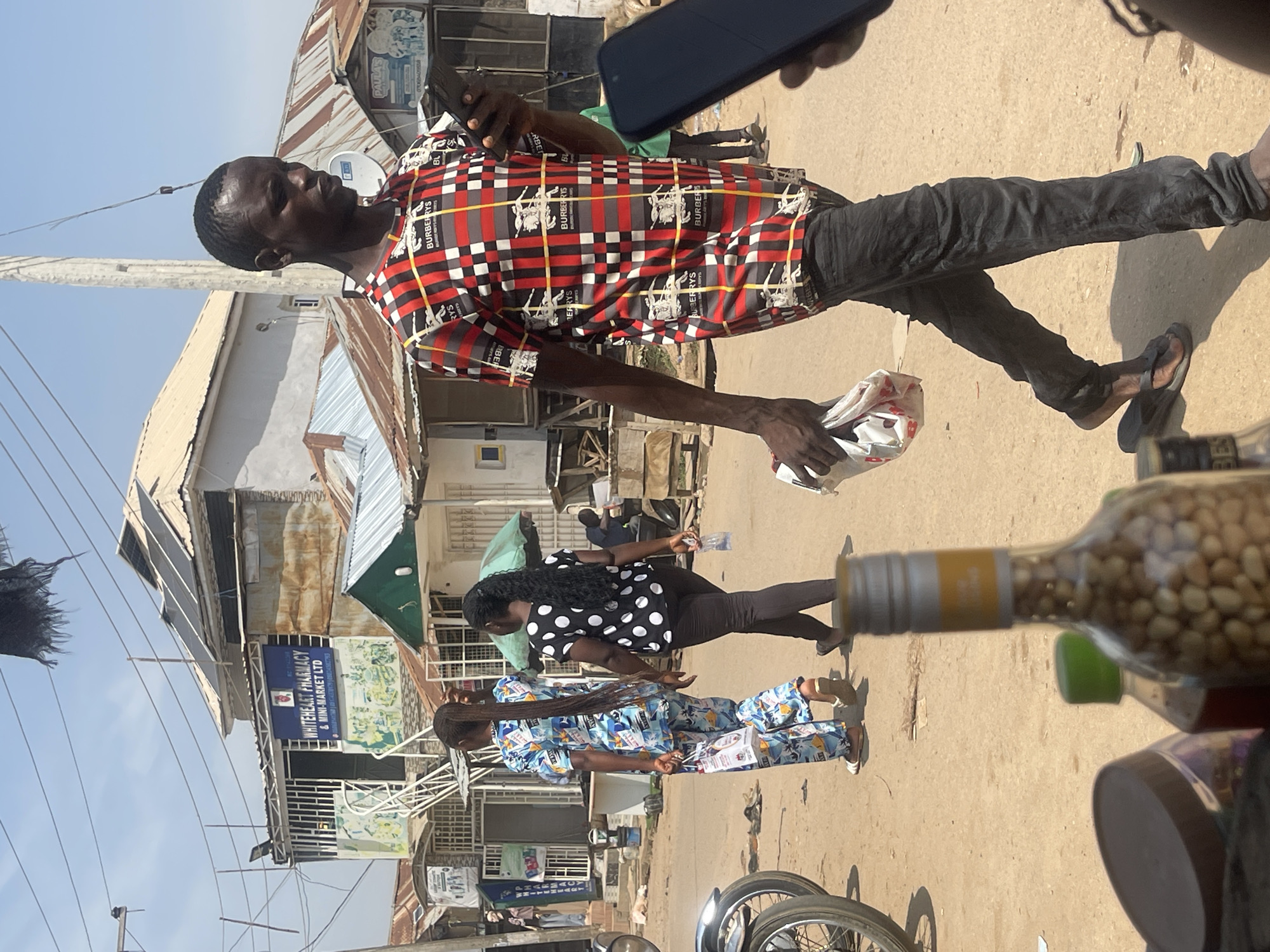 This media file depicts a street scene in Abuja, Nigeria, likely during the daytime, given the bright sunlight casting shadows. The primary focus is a man in a red, black, and white checkered shirt with a Burberry pattern, holding a plastic bag. He is walking on a dirt or paved street lined with buildings that appear to be shops or market stalls.
Behind him, a woman with long, dark hair is wearing a black and white polka dot shirt and dark pants. Another woman in the foreground is wearing a blue and white patterned outfit. Several objects are visible, including a glass bottle filled with nuts, a bicycle, and various signs. One prominent sign reads "WHITEHEART PHARMACY & MINI-MARKET LTD." Another partially visible sign says "PAULA'S." The overall impression is of a busy, everyday street scene in an urban environment. There are no explicit emotional cues, but the presence of multiple people suggests daily commerce and movement.
