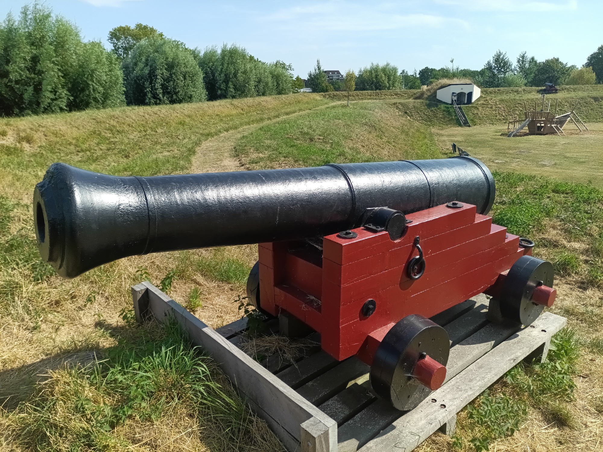 The image shows a historic black cannon mounted on a red wooden carriage with four black wheels. The cannon is positioned on a wooden platform in a grassy outdoor area. In the background, there are rolling hills covered in grass and trees, with a clear blue sky above. A small, white building with a grass-covered roof is visible on one of the hills, along with a wooden playground structure. The scene appears to be a historical site or park. Based on the context, this image is likely from the Brill fortress in the Netherlands, a location known for its historical significance and preserved fortifications.