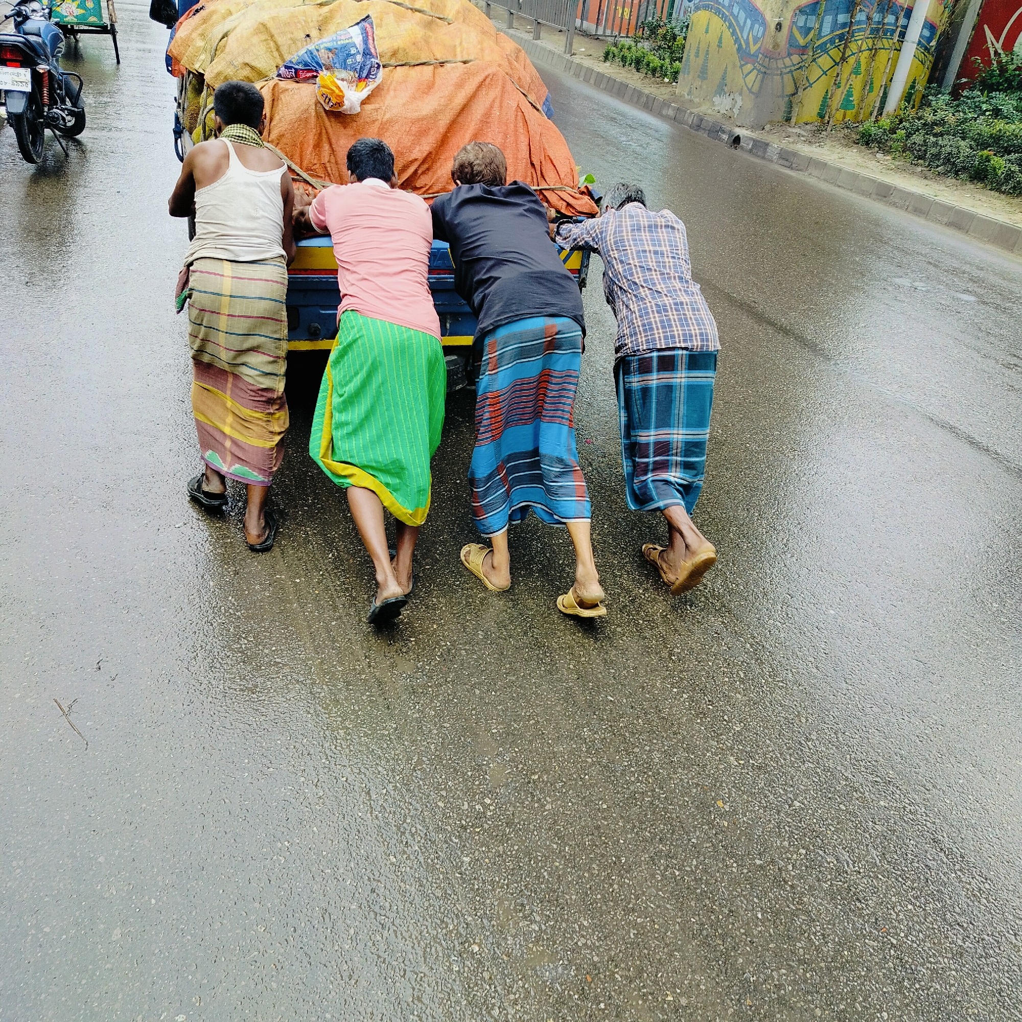 In Dhaka, Bangladesh, four men are pushing a heavily laden cart on a wet asphalt road. The cart is covered with an orange tarp, and a smaller blue tarp is visible underneath. One man in a white tank top and a striped lungi pushes from the front left. Next to him, a man in a pink t-shirt and a green lungi also pushes. Behind them, a man in a black t-shirt and a blue and red striped lungi, and a man in a plaid shirt and a blue checkered lungi push from the right. The road is wet, indicating it may be raining or has recently rained. In the background, a motorcycle is parked on the left side of the road, and a colorful mural is visible on a wall to the right. The overall scene depicts manual labor and transportation in an urban environment.