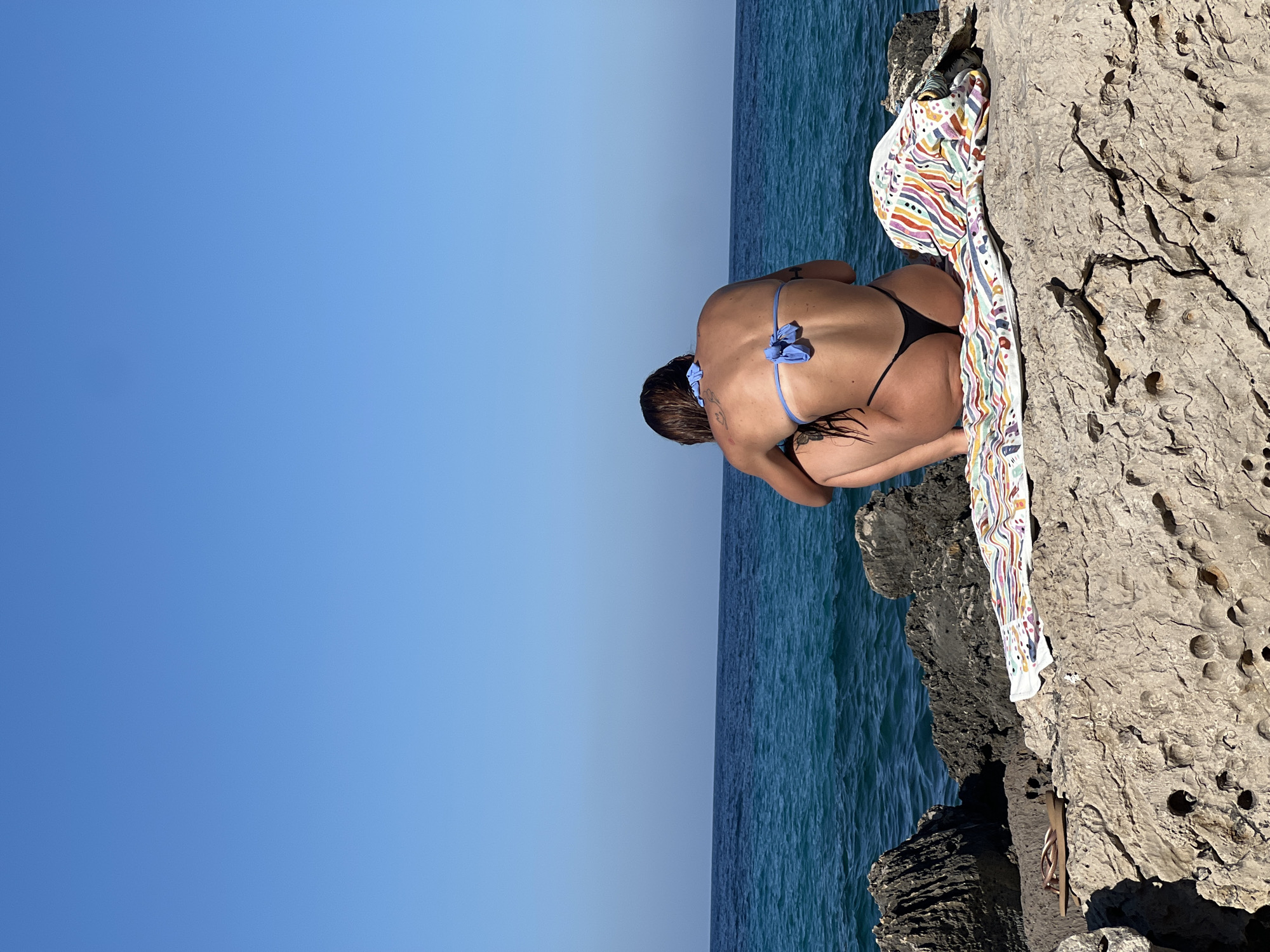 This media file shows a woman from behind, crouching on a rocky outcrop by the sea. She is wearing a blue bikini and a black thong. Her hair is dark and pulled back. She appears to be looking down at the water or the rocks. A colorful, patterned towel is draped over the rocks next to her, and a pair of flip-flops is also visible on the rocks. The sea is a deep blue, and the sky is a clear, bright blue, suggesting a sunny day. The rocks are rough and textured, with some holes and crevices. The image is oriented vertically, with the sky occupying the left side and the sea and rocks on the right. The scene evokes a sense of peace and solitude, as if the woman is contemplating the natural beauty around her. The bright sunlight and clear blue sky indicate it's likely daytime, possibly late morning or afternoon. The rough, rocky terrain and the turquoise waters suggest a coastal or seaside location.