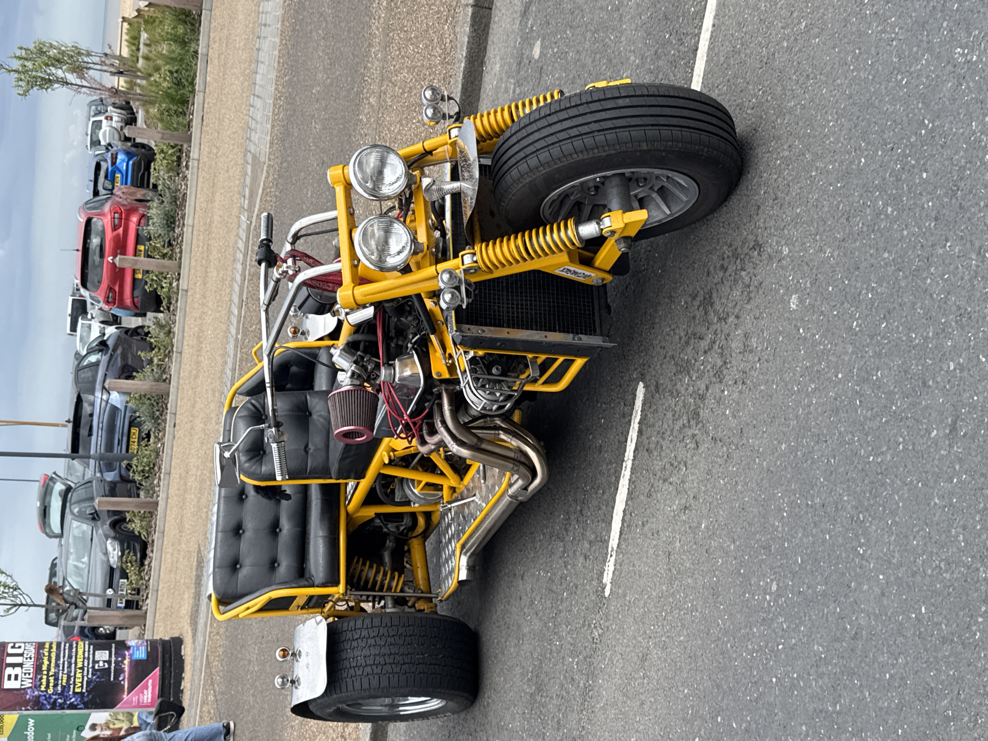 The image shows a yellow trike parked in Great Yarmouth, United Kingdom. The trike has a black, quilted seat and chrome exhaust pipes. It has two headlights at the front and a single tire at the rear. The front tire has yellow suspension. In the background, there are several parked cars and a sign advertising "Big Wednesdays." A person is visible in the lower left corner of the image. The setting appears to be an outdoor parking lot.