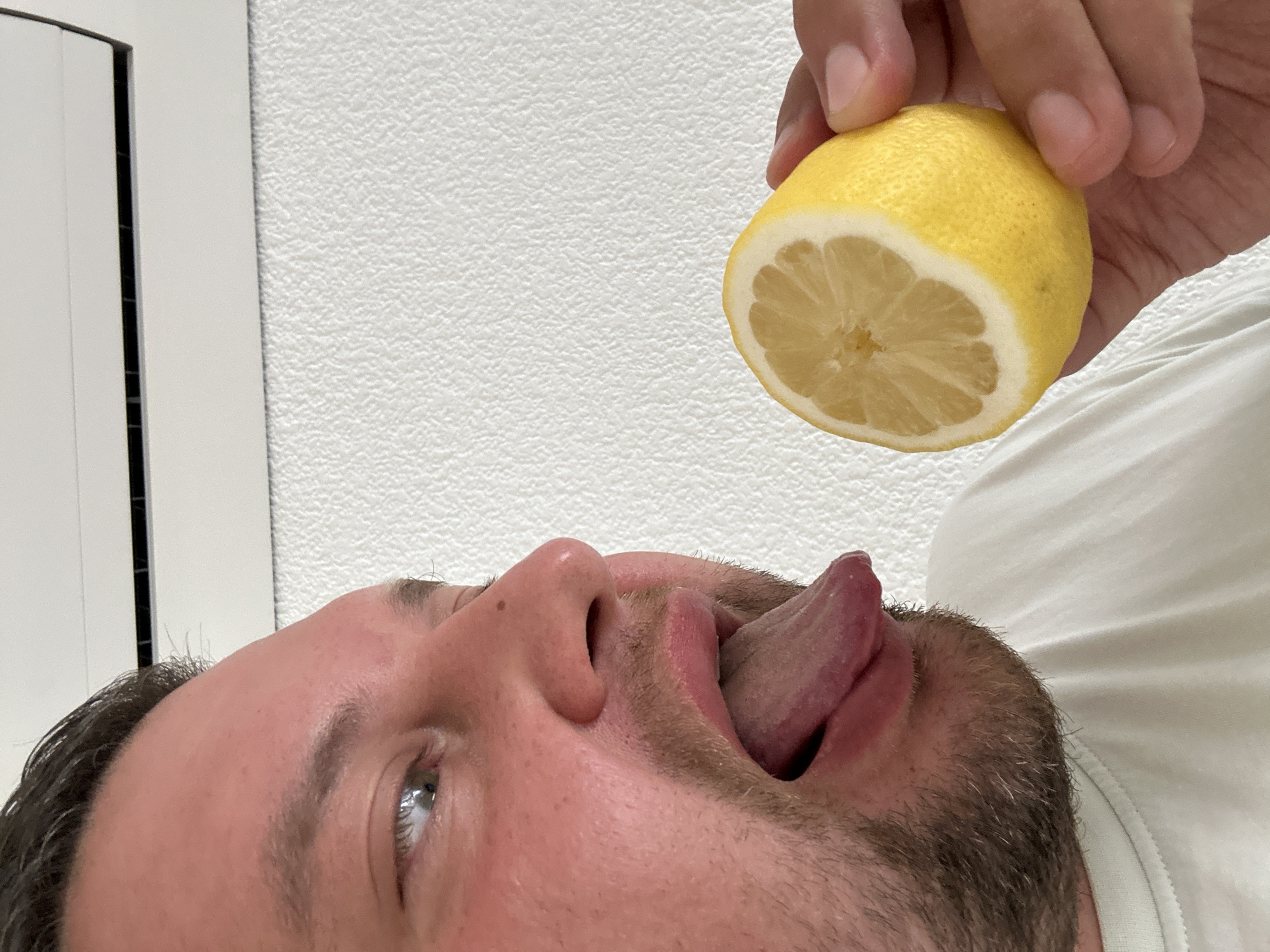 A man is lying on his back with his mouth open and tongue extended, facing upwards. A hand holds half a lemon above his mouth. The background consists of a white wall with a textured surface and a partial view of a white door or cabinet. The scene appears to be indoors. The location in Sant Feliu de Guíxols, Spain is not discernible from the image.