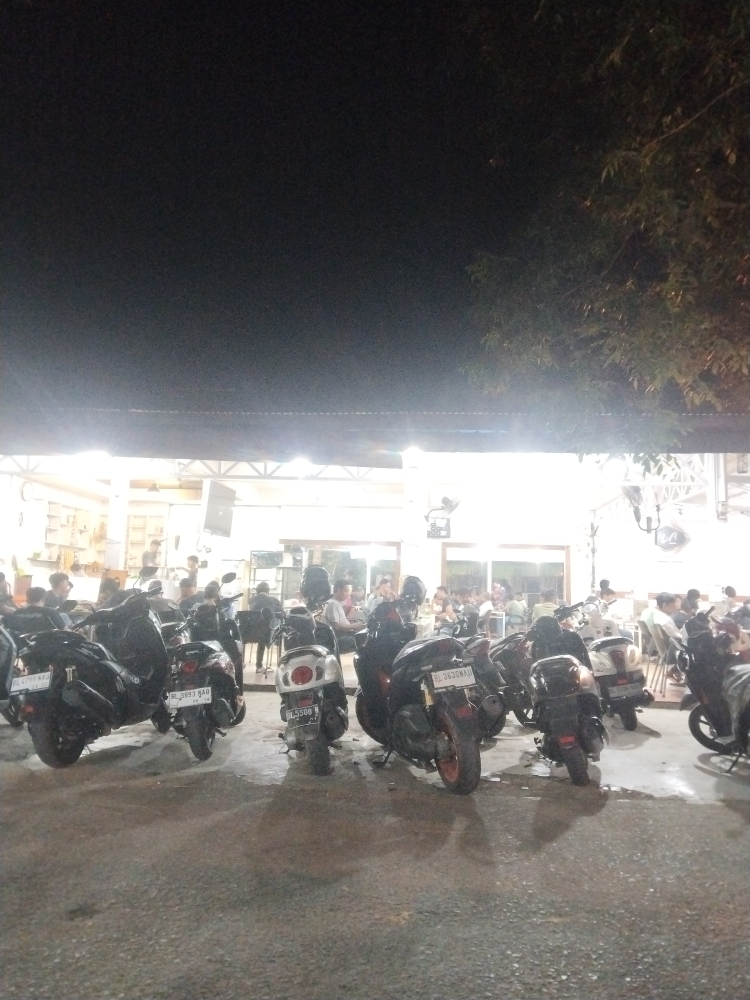 The image depicts an outdoor dining area in Lhokseumawe, Indonesia, at night. Several motorbikes are parked in front of the restaurant. Patrons are seated at tables within the brightly lit establishment. A building with a light-colored roof and interior walls is visible. The environment is dark beyond the illuminated restaurant area. Tree branches are visible in the upper right of the image.