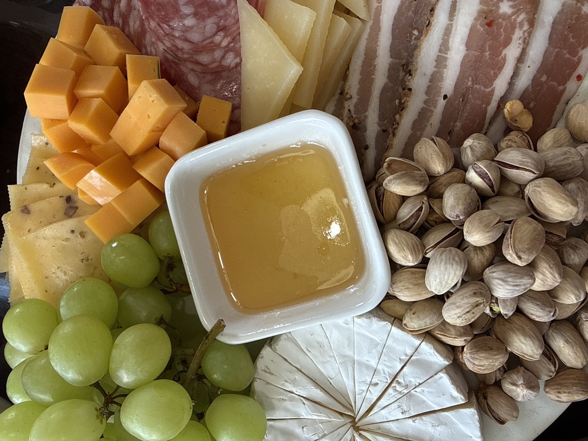 A high-angle, close-up shot depicts a charcuterie board. The board contains cubes of cheddar cheese, sliced parmesan, salami, cured meat, a small bowl of honey, green grapes, brie, and pistachios. There is no evidence to support this image being located in the city of Chaikovsky, Russia. There are no people, actions, or interactions visible.