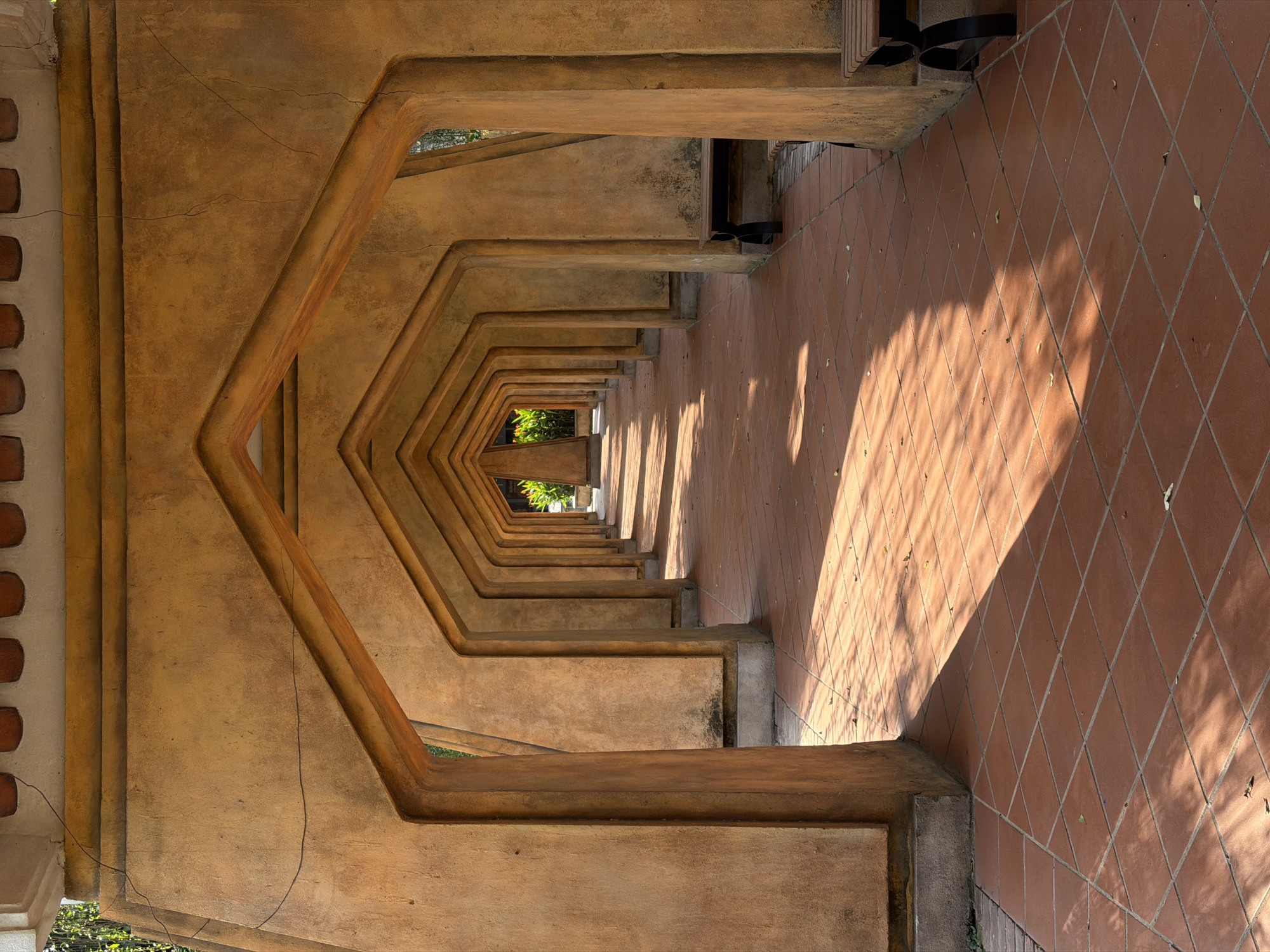 An eye-level, medium shot presents a symmetrical tunnel structure within what is likely a building in Đà Nẵng, Vietnam. The tunnel is defined by a series of repeating hexagonal frames, constructed from textured beige or tan concrete, which recede into the distance. The floor is composed of terracotta-colored tiles laid in a diamond pattern. Sunlight casts elongated shadows across the tiles, adding depth and dimension to the scene. A glimpse of green foliage is visible at the tunnel's far end, suggesting an opening to the outside. At the upper right portion of the image, the lower portion of a person, possibly wearing black clothing, is visible. The composition emphasizes the perspective and architectural details of the tunnel.