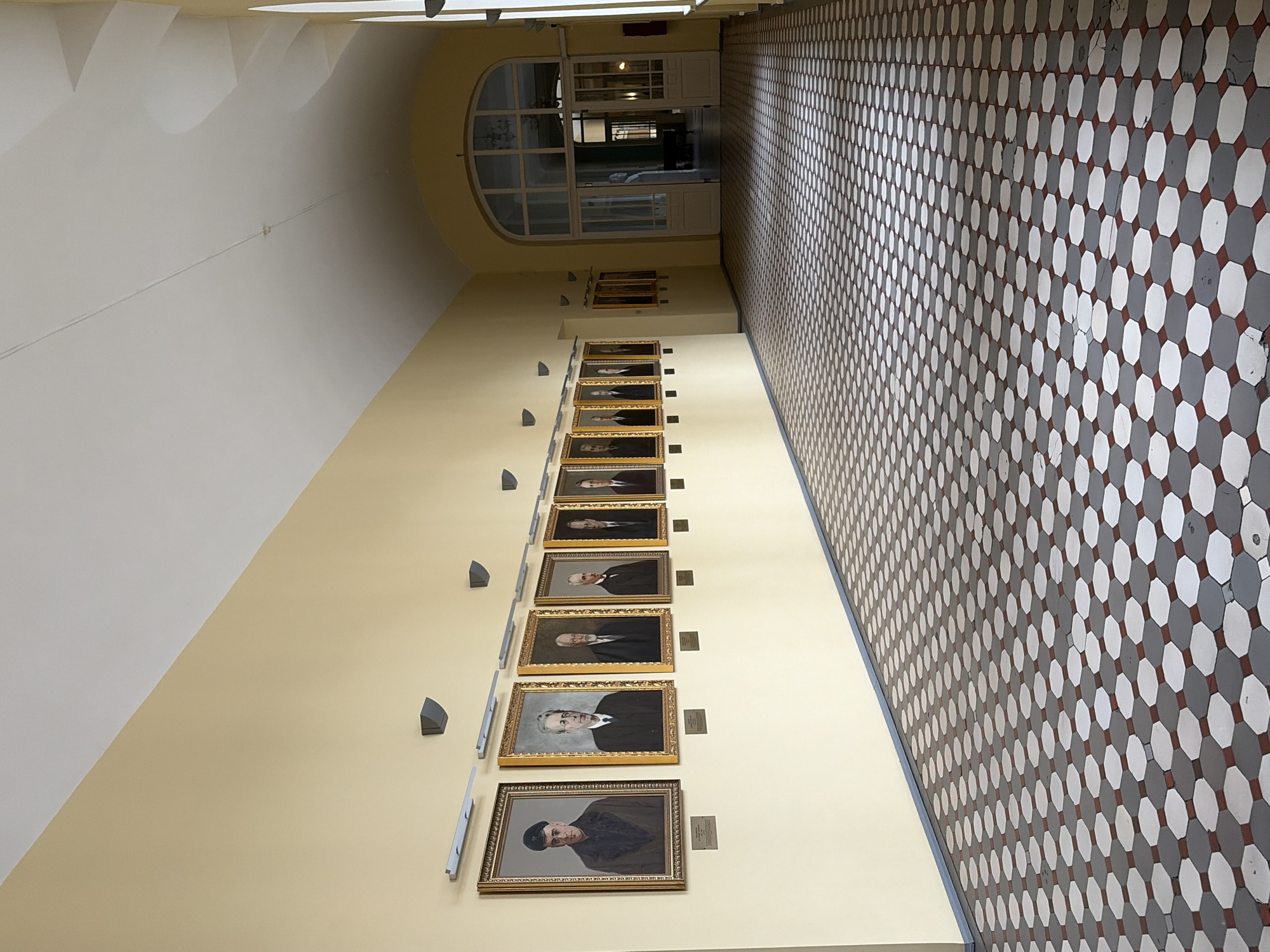 The image depicts a long, brightly lit hallway with a high, vaulted ceiling. The walls are painted a pale yellow. Along the left wall, a series of framed portraits of formally dressed men are hung at regular intervals, each with a small plaque beneath it. The floor is tiled with a repeating pattern of octagons in shades of white, grey, and red. The hallway leads to a large arched doorway with glass panels and double doors, suggesting an entrance to another part of the building. The overall impression is one of institutional grandeur. The photograph was taken in the vestibule of the Smolny Institute in Saint Petersburg, Russia.